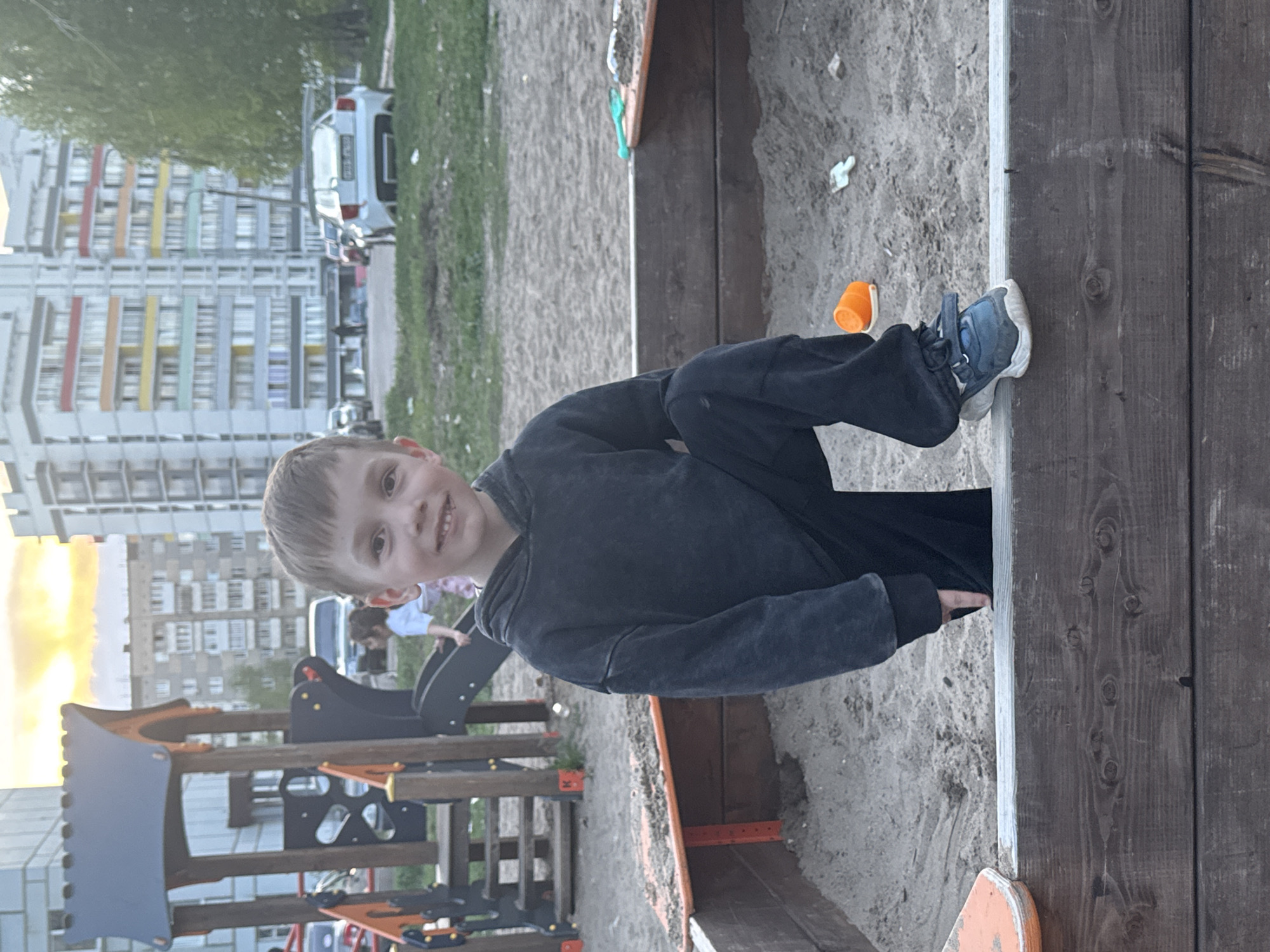 A young boy with short blond hair, wearing a dark-colored sweatshirt and pants, is standing next to a wooden sandbox in a playground. He is smiling and looking at the camera. His left foot, which is clad in a blue sneaker, is touching the edge of the sandbox. Near his foot is a small, orange plastic bucket. The playground has other equipment, including a climbing structure and a slide. In the background, there is a residential building with multi-colored balconies and a parked white car. The location is in Chaikovsky, Russia.