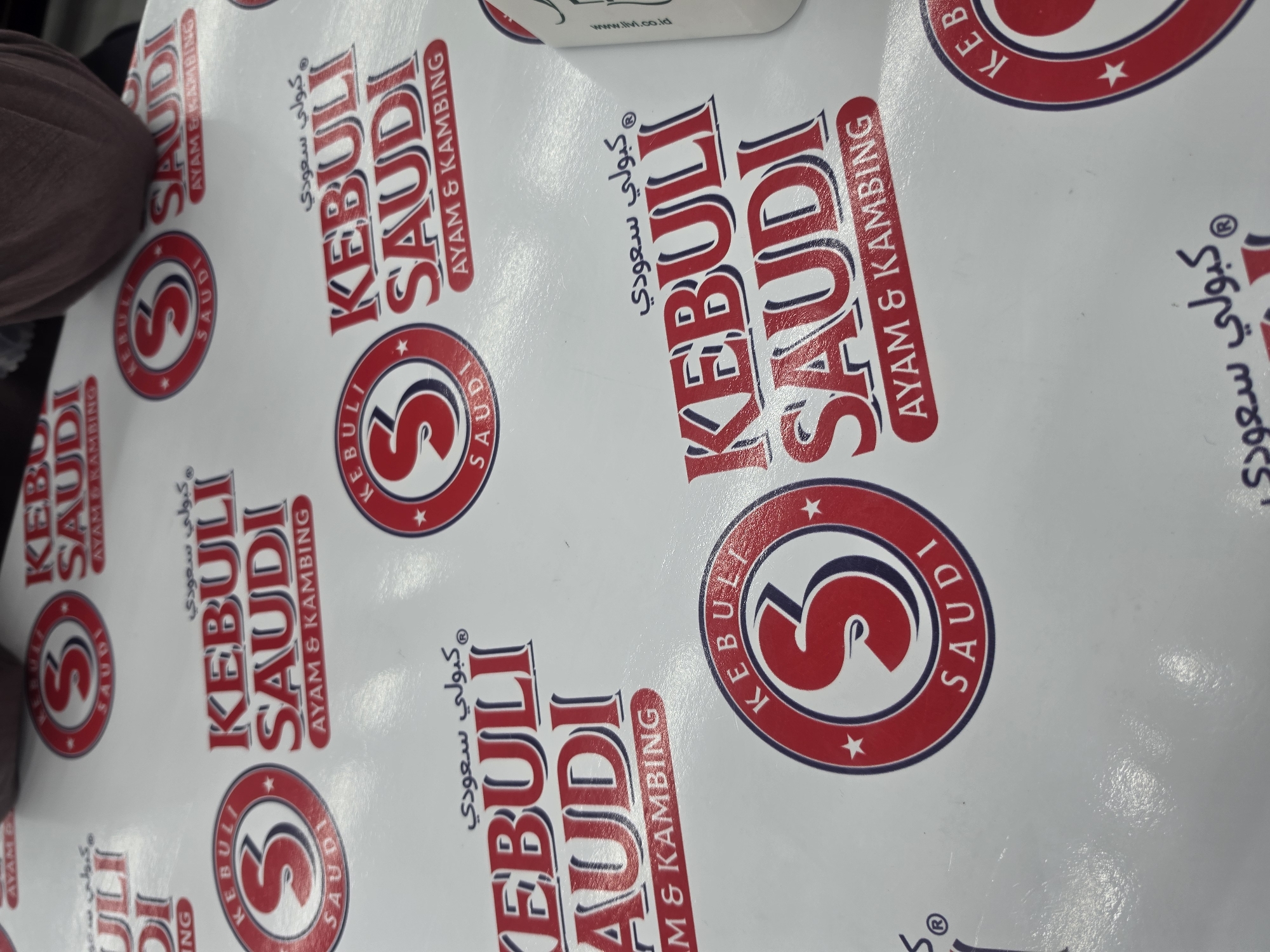 This media file showcases a close-up, overhead view of a white paper tablecloth patterned with repeating logos. The logos feature the brand name "KEBULI SAUDI" in large, stylized red and blue letters, with "AYAM & KAMBING" written below. Each main logo is also encircled by a circular emblem containing the letter "S" within a red and blue border, along with smaller instances of the brand name. Arabic script, reading "كُبولي سعودي" (Kebuli Saudi), is also incorporated into the design.
The tablecloth appears to be laid out on a surface, possibly a table, and is likely intended for food service, given the brand's name suggesting chicken and goat dishes. The lighting suggests an indoor setting, possibly a restaurant or a home dining area. There are no people or specific activities actively taking place within the frame, but the context implies preparation for or engagement in a meal. The image is cropped in a way that suggests the tablecloth might be larger than what is fully visible. The orientation of the tablecloth is slightly askew, as if the photographer is adjusting their position. The text on the tablecloth provides the brand name and type of cuisine, hinting at a Saudi Arabian-inspired dish. There are also some faint and partially visible website addresses at the very top, such as "www.lti.co.id".