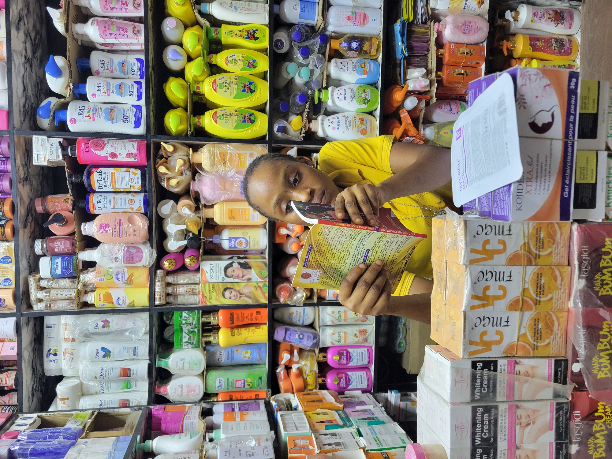This vertical medium shot shows a young woman in a yellow t-shirt intently reading a book, surrounded by a densely packed array of personal care and cosmetic products. The scene is an indoor retail setting, likely a shop or market stall in Onitsha, Nigeria, with products stacked on shelves and in cardboard boxes.
The shelves are filled with numerous bottles of lotions, body washes, shampoos, and jars of creams, as well as boxed soaps and other beauty items. The woman holds an open book titled "ENJOYING SEX IN MARRIAGE BY BRO. ASIKA STANLEY CHINEDU," with a section labeled "ABOUT THE AUTHOR" visible on the page she is reading. Her expression is focused, indicating deep concentration on the book's content.
Prominently visible brands and product types include E45 Junior, Dr Teal's, Dove, Clere, Johnson's, St. Ives, Biotin, Keto, Fayan Kou, Kojide Xtra91 (a "Gel éclaircissant pour la peau" or skin lightening gel), FMGC 'VC' soaps, and NAIJA BUMBUM creams. Other products feature descriptions such as "Whitening Cream for sensitive Areas" with "COLLAGEN & MILK QUICK WHITENING," "Baby Diary" and "Teens" ranges, "Xtract Gold" products, and "Snail 94" cream. The abundance and variety of products suggest a busy commercial environment.