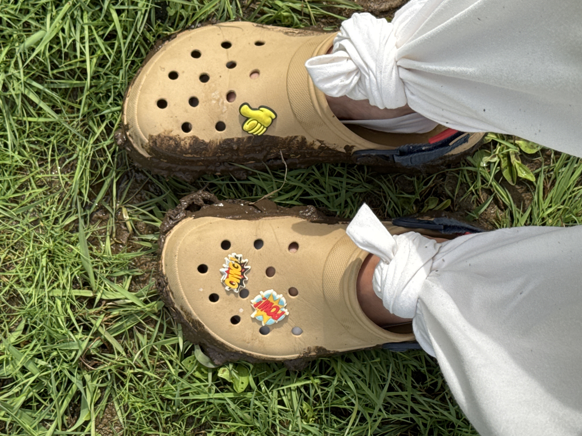 The image shows a view from above of two tan Crocs, covered in mud, on green grass. The Crocs are adorned with novelty charms in the holes. The right Croc has a yellow cartoon hand charm. The left Croc has an "OMG!" and "POW!" comic book-style charm. White pants are tied at the ankles and are tucked into the Crocs. The background consists of green grass, with some patches of dirt and mud. The location of Nozhovka, Russia, is not relevant as it is not visible in the image.