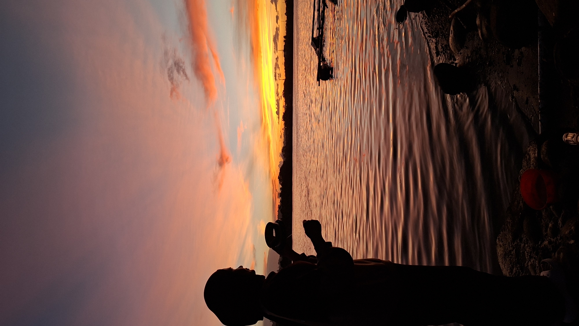The image depicts a serene sunset scene by the water, likely in Lhokseumawe, Indonesia. The sky is ablaze with vibrant hues of orange, yellow, and pink from the setting sun, casting a warm glow on the rippling water. The reflection of the sky creates a beautiful, textured pattern on the water's surface.
In the foreground, a person is silhouetted against the dramatic sky and water. They are facing away from the camera, looking out at the sunset. Their posture suggests a moment of quiet contemplation or enjoyment of the natural spectacle. The silhouette is dark, making it difficult to discern specific features, but it appears to be an adult, possibly sitting or standing by the water's edge.
The setting appears to be a shoreline, with dark, earthy textures visible to the right of the person. There are also hints of objects on the shore, including a red bucket and some rocks, suggesting a natural or perhaps a slightly developed waterfront. In the distance, the horizon is marked by a dark silhouette of trees or land, separating the sky and water. A small boat or structure is faintly visible on the water in the mid-ground.
The time of day is clearly sunset, as indicated by the dramatic colors in the sky and the long shadows. The weather appears to be clear, allowing for an unobstructed view of the sunset. The overall mood evoked by the image is one of peace, tranquility, and awe inspired by the beauty of nature. There is no visible text in the image.