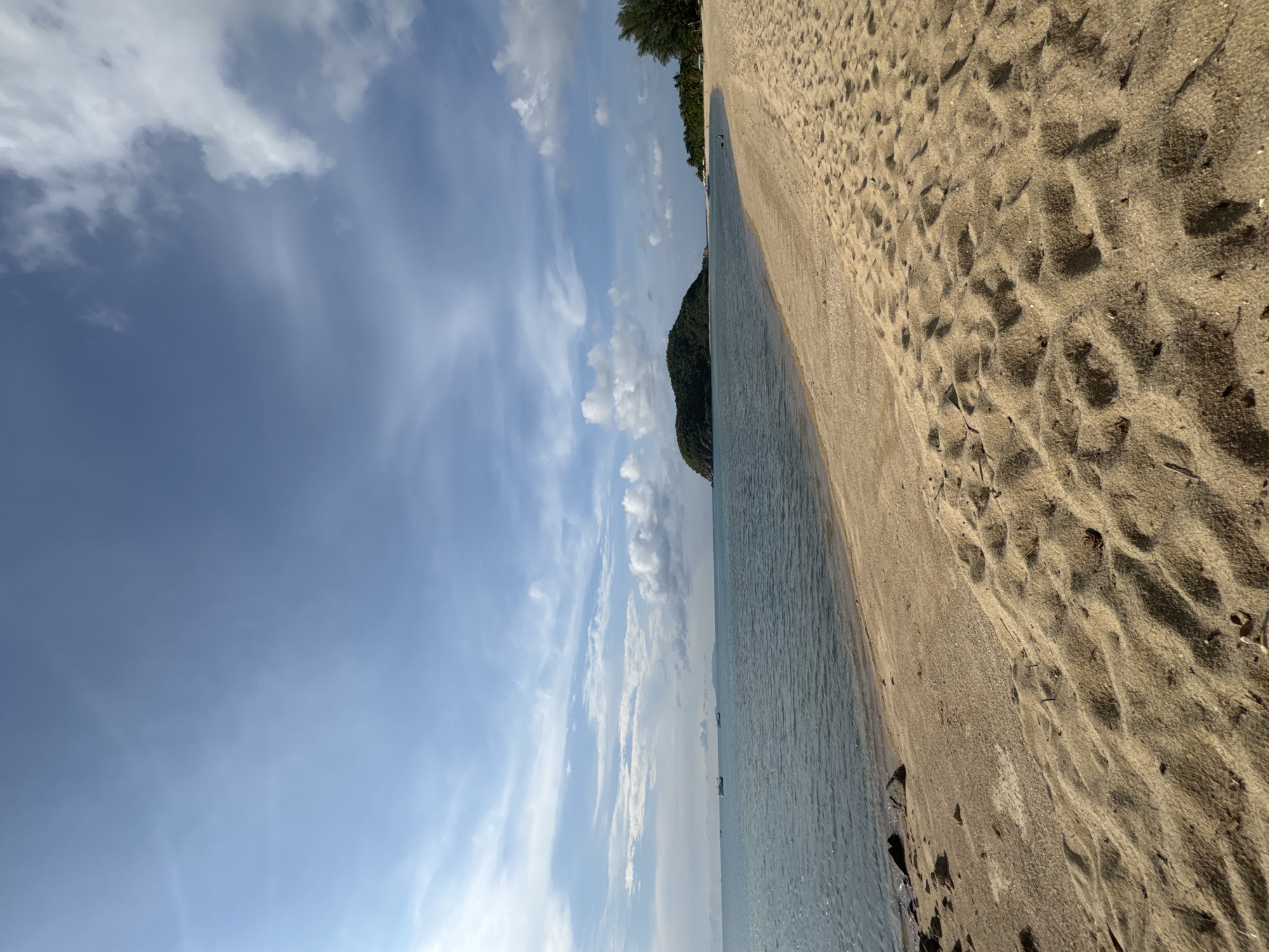 This is a landscape photograph of a beach located in Baan Wang Ta Kien, Thailand. The image has been rotated. The right side shows a sandy beach with the imprint of waves. The left side depicts water and sky with clouds. The sky is blue and partially filled with clouds. There is a tree covered land feature stretching into the ocean. The photograph captures a calm, natural environment. There are no people or animals present.