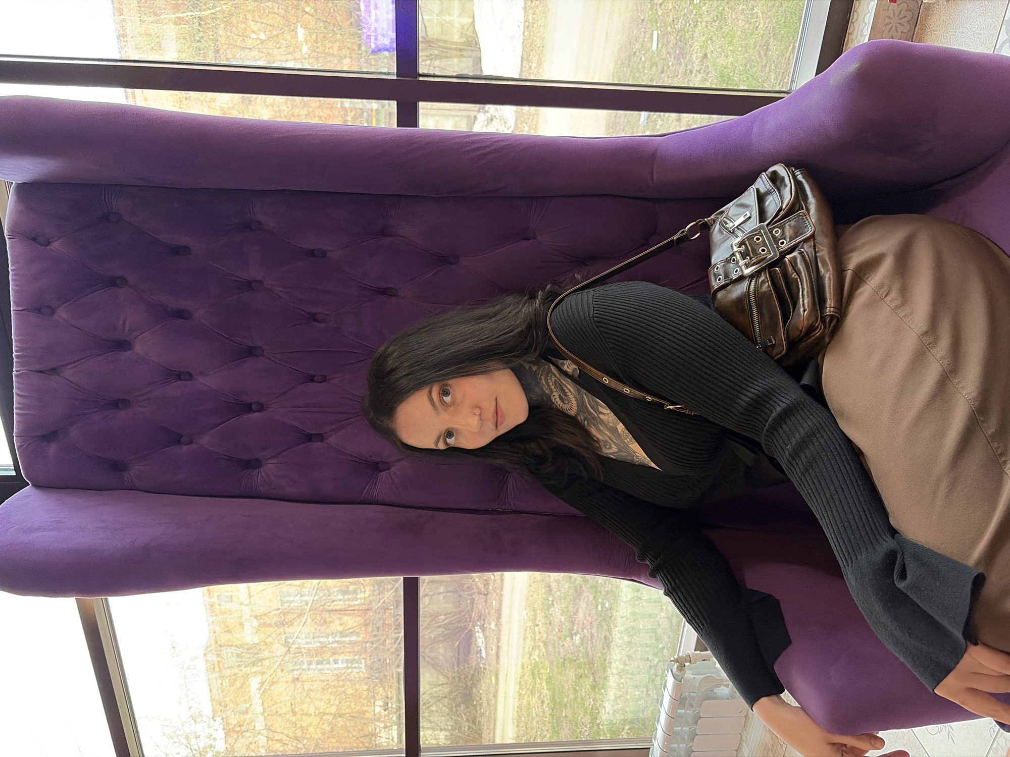 The image, rotated 90 degrees counter-clockwise, depicts a young woman seated in a large, plush, purple upholstered armchair or sofa. The chair features a tufted backrest and smooth seat. The woman has long, dark hair, light-colored eyes, and is looking directly towards the camera with a neutral expression. She wears a black, ribbed long-sleeved top, possibly a turtleneck, and what appear to be brown or khaki wide-leg trousers. Visible tattoos extend from her neck/chest area down her arms. A dark brown leather handbag with buckle and stud details rests on the armrest of the chair, near her hip.
The setting is indoors, with the woman positioned beside a large window that offers a view of the outdoors. Through the window, bare trees, patches of green and brown ground cover, and a distant building with an orange-brown façade are visible. The lighting suggests it is daytime, possibly overcast or early spring/late autumn, given the appearance of the trees and ground. The overall atmosphere appears calm and relaxed. No visible text is present in the image. The location is Chaikovsky, Russia.