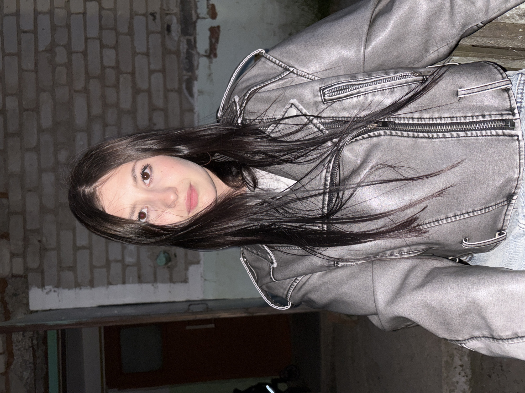 A young woman with long, dark hair is wearing a gray jacket. The background includes a brick wall, a door, and what appears to be a city street. The image was taken in an urban environment.