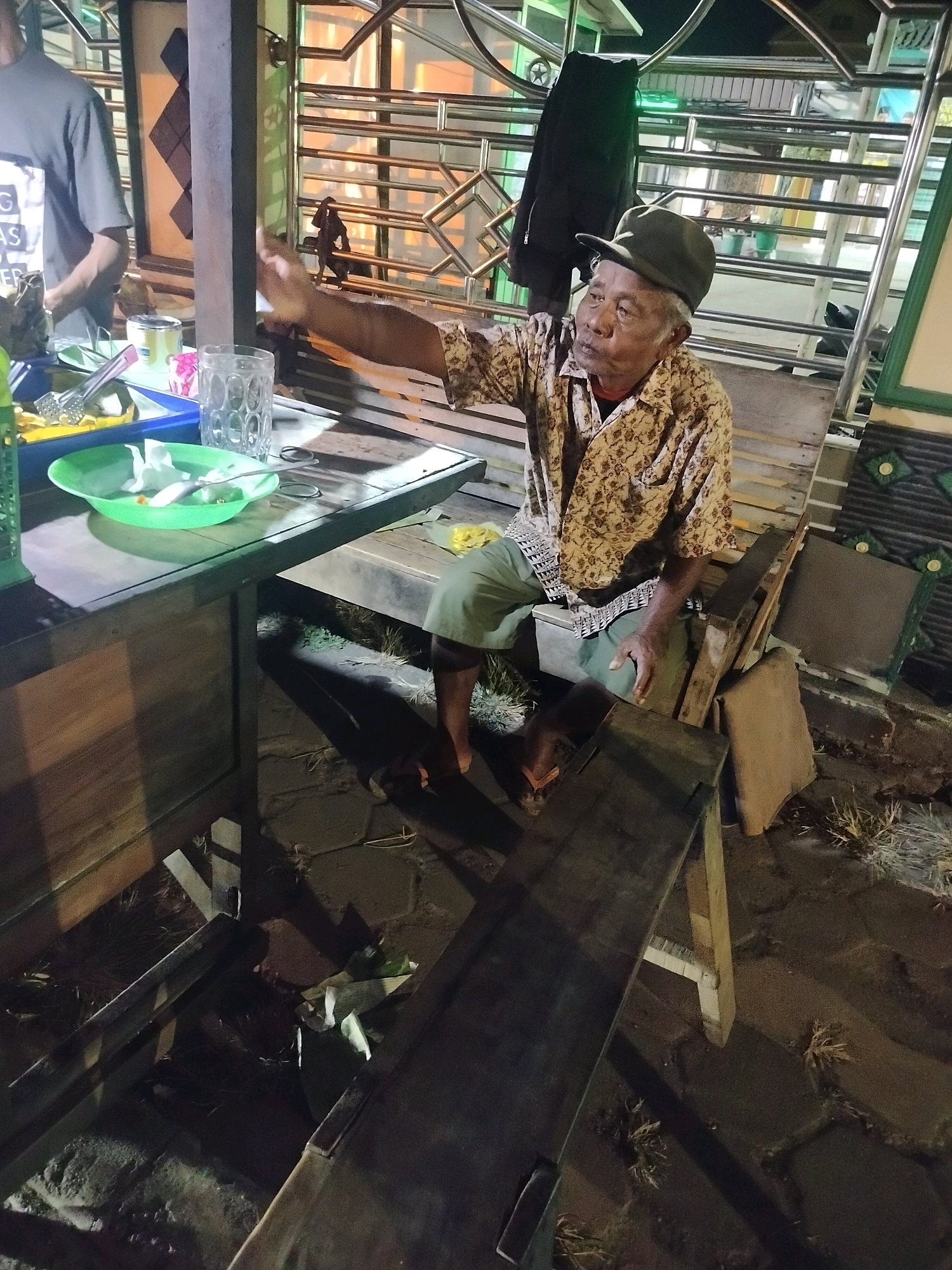 In the city of Kroya, Indonesia, an elderly man wearing a patterned shirt and a dark cap sits on a wooden bench. He is looking towards his left and has his left arm extended. In front of him is a table with a blue plastic tray containing green bowls, one of which holds crumpled white paper. There are also glasses and other items on the table. A portion of another person, wearing a dark grey t-shirt, is visible to the left of the frame. The scene appears to be outdoors, with cobblestone ground and some vegetation visible. The lighting suggests it is either dusk or nighttime, with artificial lights casting shadows.