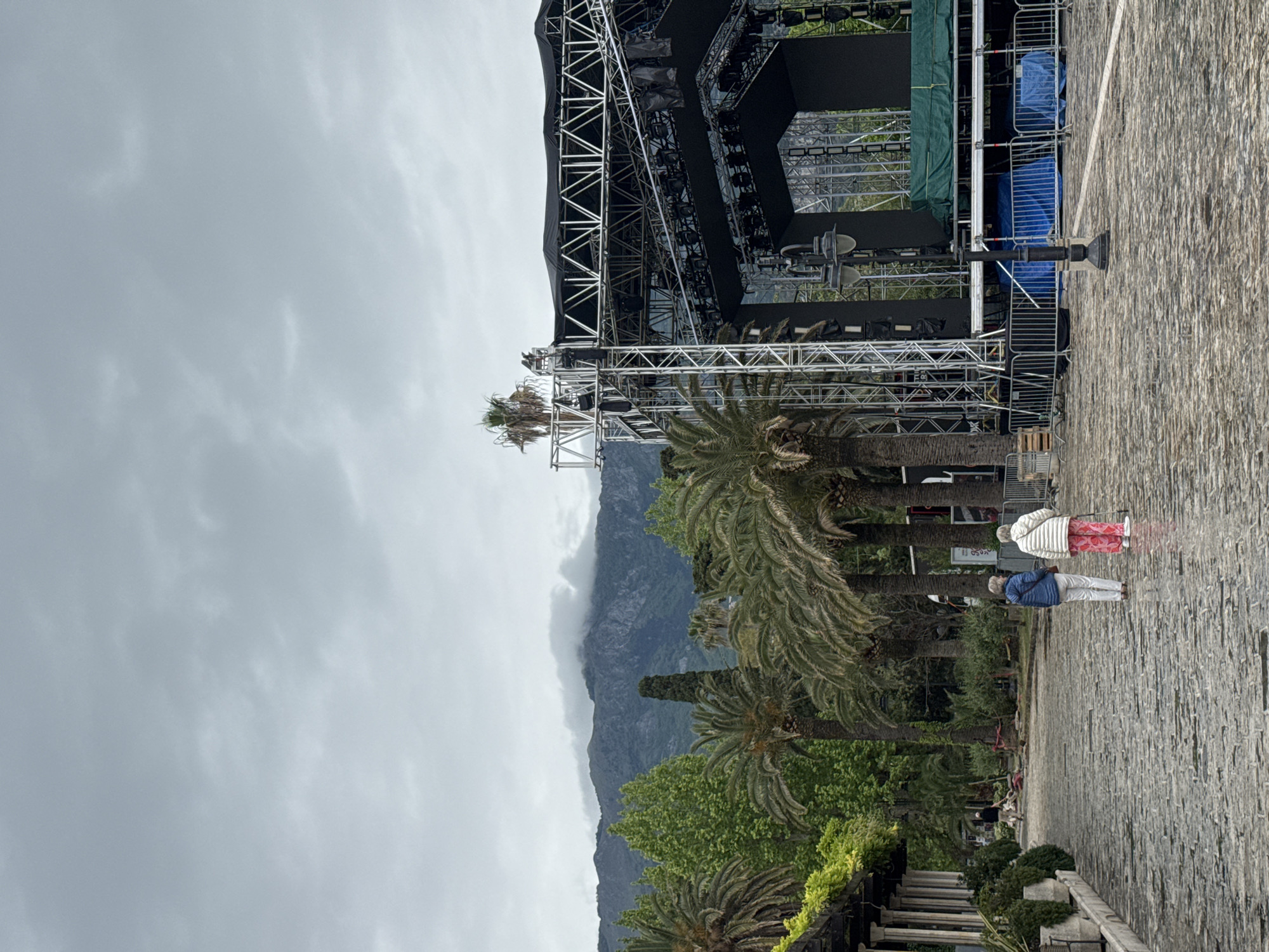 The scene is in Budva, Montenegro, on a cobblestone walkway. Two elderly individuals are walking; one wears a blue jacket and white pants, while the other wears a white jacket and red patterned pants. A large stage structure is set up nearby. Palm trees and other greenery are present. In the background, a mountain is partially obscured by clouds. The sky is overcast.