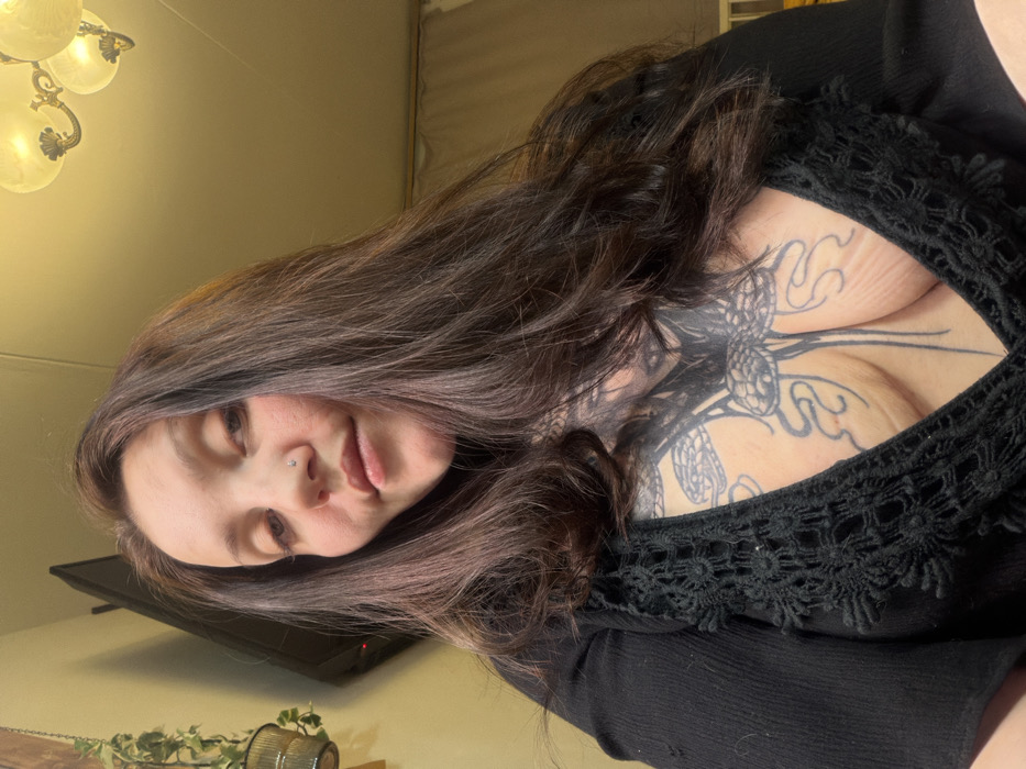 A woman poses for a selfie in a dark, low-cut top with lace detailing. She has long, dark brown hair and a nose piercing. A large tattoo covers the upper part of her chest. In the background, a television and a decorative plant can be seen. The ceiling has a chandelier.