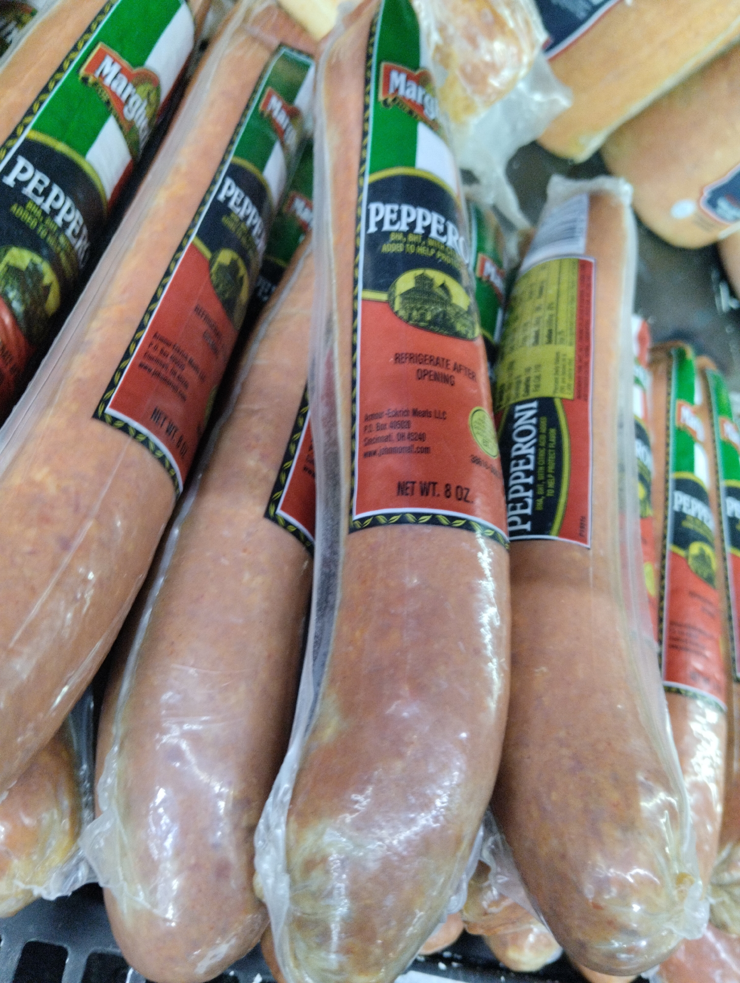 This is a close-up shot of several packages of "Margherita Pepperoni," each encased in clear plastic wrap. The packages are stacked, and the focus is on the labels. The labels display the brand name "Margherita" in a green, white, and gold design, along with the word "PEPPERONI" in bold, black letters. Below the brand name, there's an image of a building. The label also includes text indicating that the pepperoni should be refrigerated after opening and lists the manufacturer as "Armour-Eckrich Meats LLC" with a P.O. Box address in Cincinnati, OH. The net weight is stated as 8 oz. In some packages, the word "PEPPERONI" is also present in black letters on a yellow background, mentioning "BHA, BHT, WITH CITRIC ACID ADDED TO HELP PROTECT FLAVOR." The pepperoni sausages themselves are visible through the plastic, revealing a light orange-red color. The packages are resting on a dark grey surface, possibly a shelf or rack.