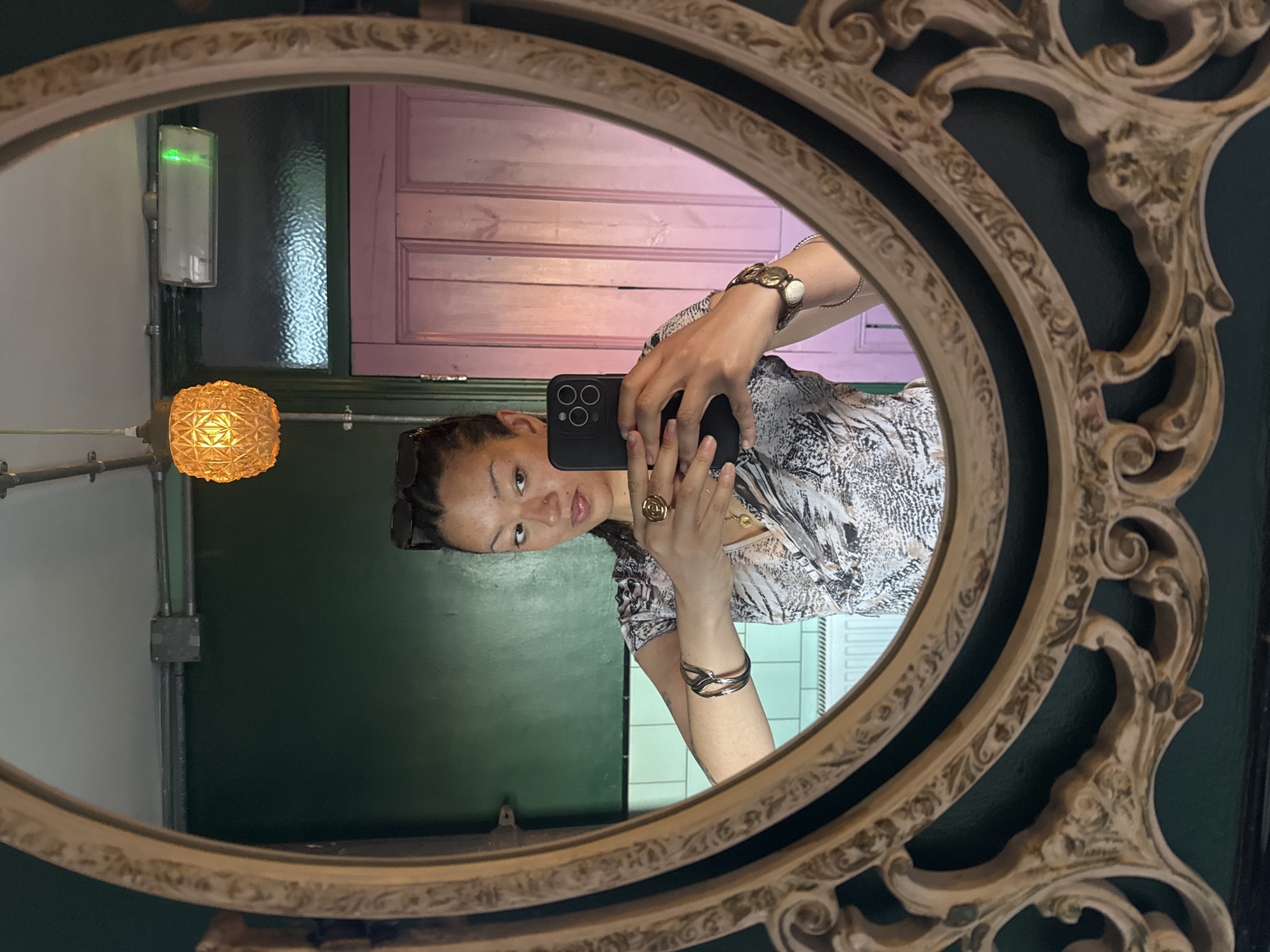 A person is reflected in an ornate, oval-shaped mirror. The reflection shows the individual holding a mobile phone and taking a photograph of themselves. The background suggests a room with green walls and a pink door, with a light fixture visible. The scene appears to be indoors. Location is London, United Kingdom.