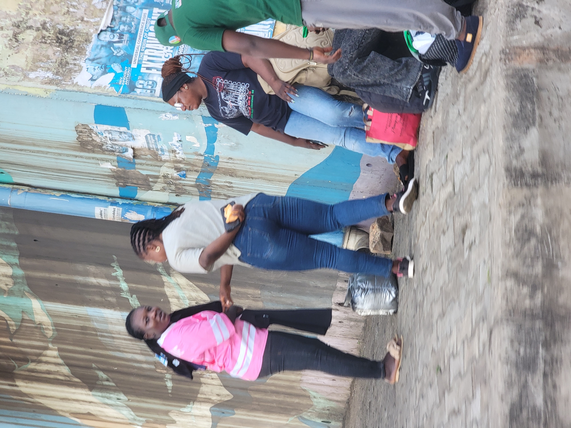 This outdoor image, captured in Jos, Nigeria, shows a casual gathering of several individuals against a corrugated metal wall. Three women are prominently featured, standing in various positions, and parts of two other people are visible.
The woman in the foreground wears a pink and white striped jacket over a black top, black pants, and light-colored sandals. Her hair is styled in dark braids pulled back. Behind her, another woman wears a light-colored t-shirt, blue jeans, and dark shoes, holding a phone. Her hair is also braided. Further back, a third woman wears a black t-shirt with a white graphic design, blue jeans, and sneakers. She has dreadlocks or braids tied back with a black headband and wears glasses. Next to her, a man in a green shirt is partially visible, along with the legs and shoes of another person. A red bag rests near the feet of the woman in the black t-shirt, and a silver/grey bag is near the feet of the woman in the white t-shirt.
The background consists of a weathered corrugated metal wall, painted with large blue and brown/grey sections. Numerous old posters and flyers are peeling off the wall, revealing layers beneath. Visible text on these posters, though worn, includes "UNIVERSAL CONFERENCE 2025," "THE REIGN OF CHRIST," and "REV. INNOCENT ANTHONY," with a specific location mentioned as "Cleveland, OH." The ground is paved with interlocking concrete blocks. The scene appears to be taking place during the daytime, under natural light, and the overall atmosphere is candid and observational. The people seem to be standing and possibly conversing, engaged in an everyday street activity.