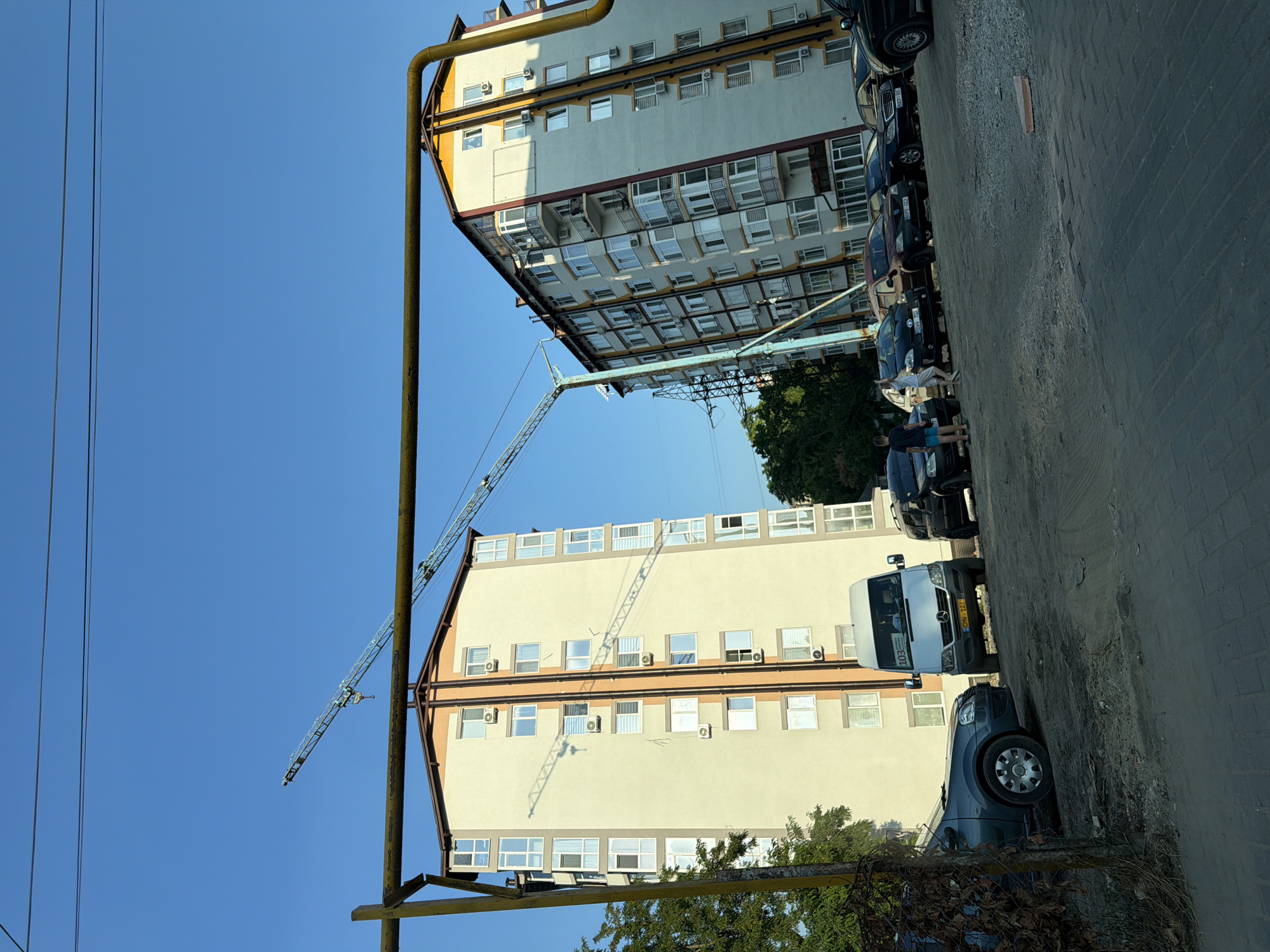 The image depicts an urban scene in Durlești, Moldova, with two multi-story apartment buildings dominating the background. A construction crane is visible in the sky, extending over the buildings. In the foreground, several vehicles are parked, including a white van with "103" displayed on its windshield, a dark sedan, and other cars. Two individuals are standing near the vehicles, appearing to interact. The ground is a mix of paved surfaces and dirt. The sky is clear and blue, suggesting a daytime scene. Trees are present on the left and right sides of the frame, bordering the buildings. A yellow pipe structure runs across the upper portion of the image, obscuring parts of the sky and crane.