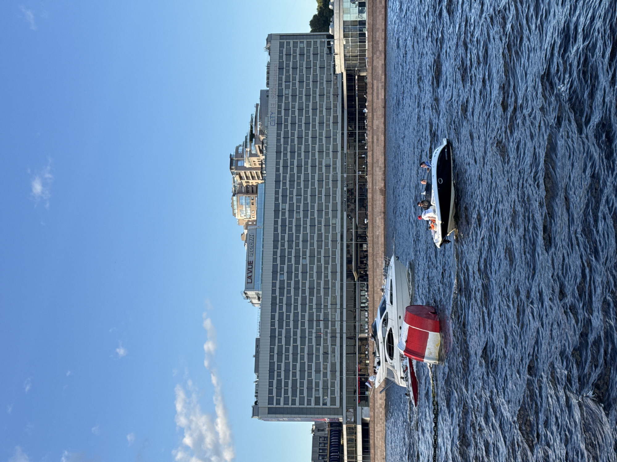The image depicts a daytime scene on the Neva River in Saint Petersburg, Russia, with a large modern building complex lining the embankment. In the foreground, the dark blue water of the river is choppy, with whitecaps visible. Two boats are present: a sleek, white motor yacht with several people on board is moving away from the viewer to the left, and a smaller, black and white speedboat with four people is closer to the right, also moving left. A red and white buoy is partially submerged in the water between the two boats, attached to a chain.
The building complex behind the river is a multi-story structure with a predominantly gray facade and numerous windows. The upper floors feature a stepped architectural design with balconies and rooftop elements. Signage on the building indicates "LA VUE РЕСТОРАН" (La Vue Restaurant) and "CITY HOTEL" with "САНКТ-ПЕТЕРБУРГ" (Saint Petersburg) beneath it. The sky above is a clear blue with a few scattered white clouds. The overall impression is a bustling urban waterfront scene on a clear day.