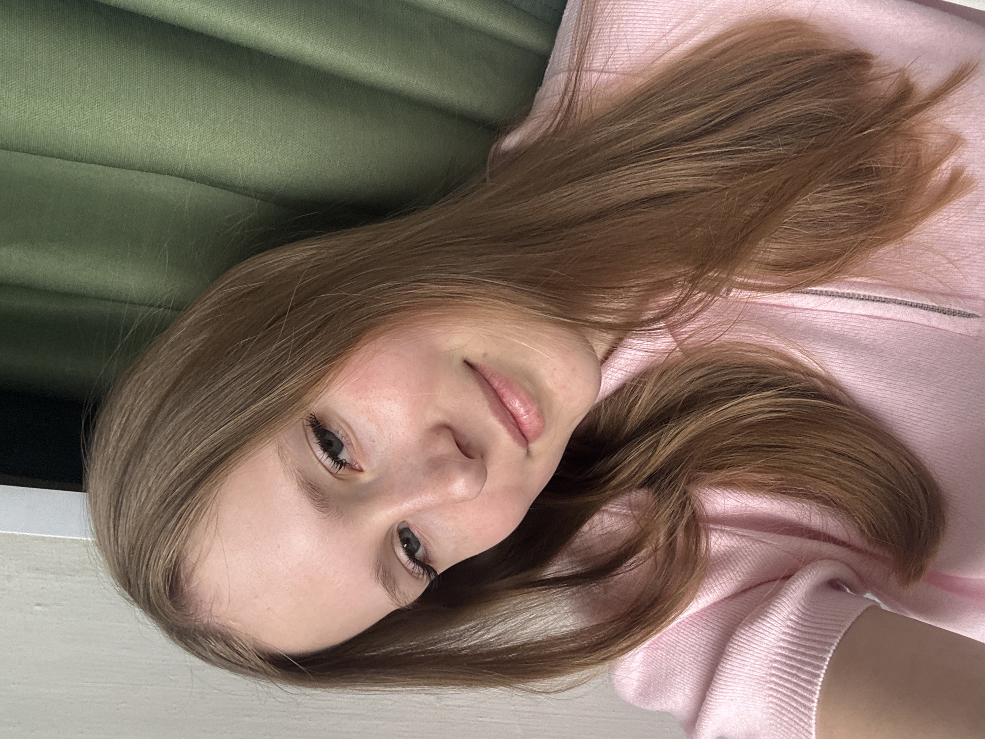 A young woman with light skin and light brown hair is lying down, with her head slightly tilted to the left. She is wearing a pink sweater. The background includes a green curtain or fabric. The specific location in the city of Chaikovsky, Russia is not discernible from the image.