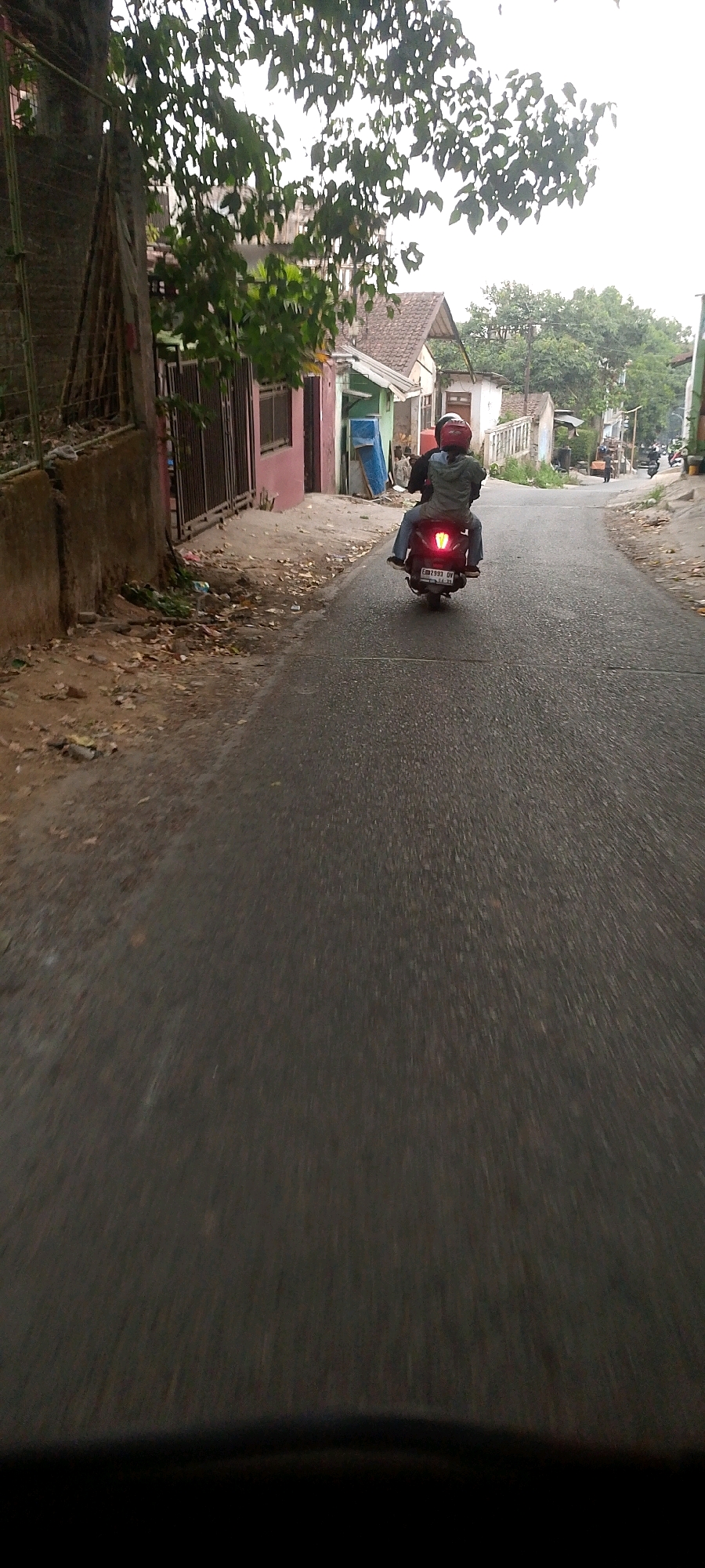 A two-person motorcycle is traveling away from the camera on a paved road in a residential area. The driver wears a red helmet, and the passenger, in a green jacket, has their arms around the driver. The motorcycle's red taillight is illuminated. The road is flanked by houses and vegetation, with a sidewalk on the left side of the road featuring some debris. The sky is overcast. The location appears to be in or around Bandung City, Indonesia, based on the context provided.