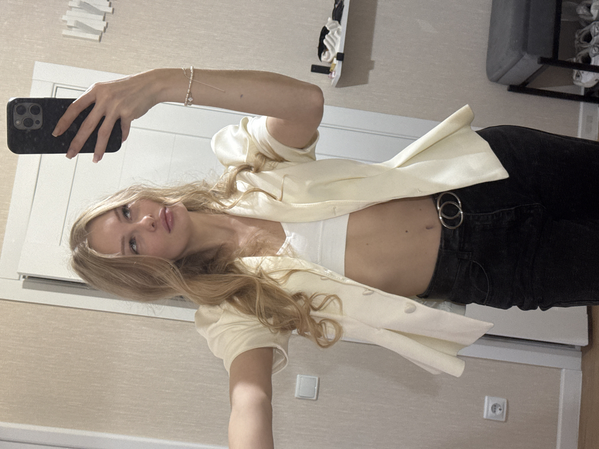A young Caucasian woman with blonde wavy hair stands in a room, taking a mirror selfie with her phone. She is wearing a cream-colored blazer over a white crop top and black jeans with a silver double-ring belt. The room appears to be a bedroom or dressing area, with beige textured wallpaper and a white door in the background. There is a power outlet on the wall to the left of the woman, and a shelf with toiletries and accessories is visible to the right. A grey upholstered stool with shoes underneath is also in the background. The lighting is soft and even, suggesting an indoor setting. There is no indication of the city of Perm, Russia, in the image.