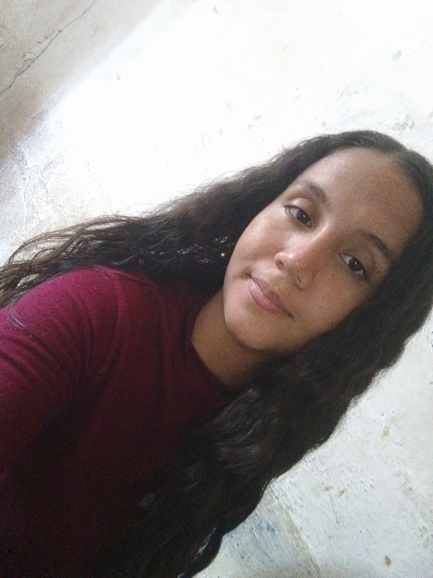A woman with long, dark hair is posing for a selfie indoors. She is wearing a maroon shirt. The background appears to be a roughly textured wall, possibly painted white. The location is not definitively identifiable as Juan Griego, Venezuela, based on the image alone. There are no other people, subjects, objects, actions, or interactions visible in the provided image.