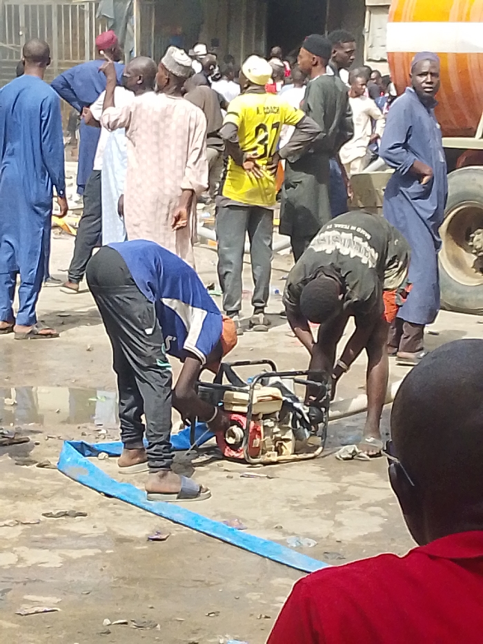 This image depicts an outdoor urban scene in Fagge C, Nigeria, featuring a crowd of approximately 15-20 individuals, predominantly adult males. In the center-midground, two men are actively engaged with a portable, gasoline-powered water pump, characterized by its red and beige engine housing and black frame. One man, wearing a blue t-shirt, dark pants, and sandals, is bent over from the left, adjusting a component of the pump. The second man, in a dark t-shirt and dark shorts or pants, is similarly bent over from the right, also interacting with the pump. A blue lay-flat hose lies on the wet, uneven ground, connected to or extending from the pump towards the left.
The surrounding area is populated by a standing crowd of men. Notable individuals include a man in a bright yellow jersey with "A. COACH 37" printed on the back, and several men in traditional Nigerian attire, including long tunics and skullcaps. On the far right, a large vehicle tire and a segment of a yellow tank are partially visible, indicating the presence of a heavy vehicle, possibly a truck or tanker. The ground is a mix of damp, dirty earth and concrete, with visible puddles and scattered debris. The scene occurs during daylight hours under bright illumination.