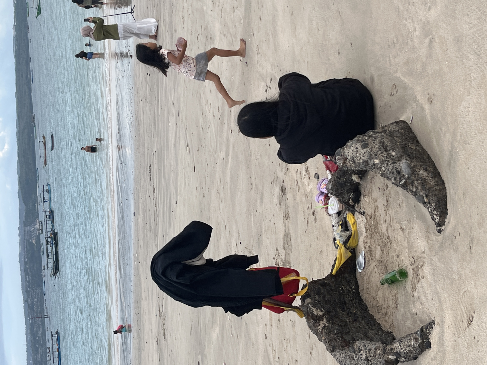 The image depicts a beach scene in Kuta, Indonesia, with the ocean and a distant shoreline visible in the background. In the foreground, a black jacket and a red bag are draped over a rock. Nearby, various items are scattered on the sand, including a green bottle, flip-flops, and plastic trash. Two individuals are present: a young girl in a patterned shirt and skirt is running across the sand, and another person is seated on the beach, facing away from the camera. In the water, several people are swimming or standing, and numerous boats are anchored offshore. The sky is overcast, suggesting a cloudy day.