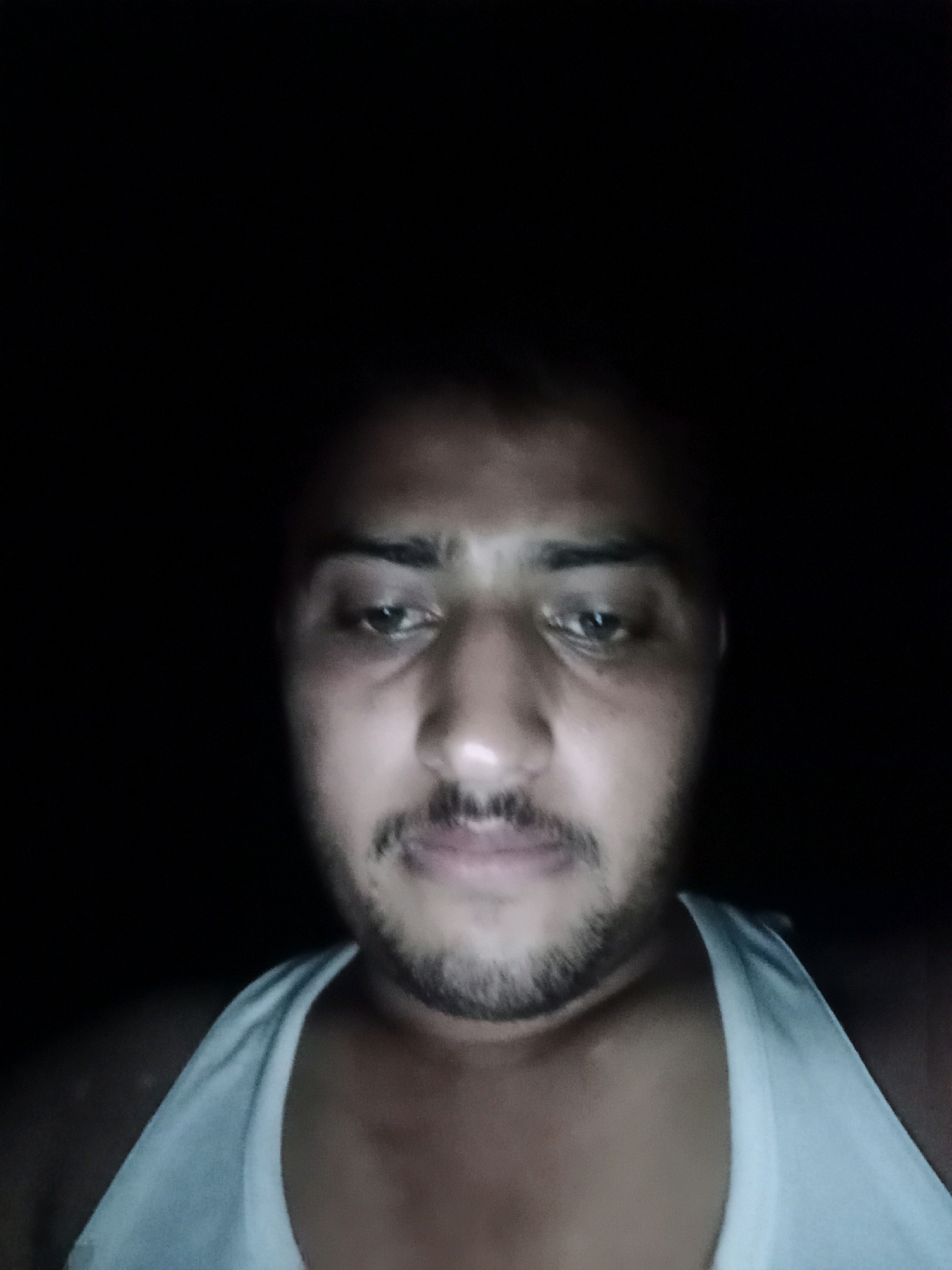 The image is a close-up, low-light selfie of a man. He is wearing a white tank top. The background is dark and out of focus, making it impossible to determine the specific location within Chak 234 A, Pakistan, or any environmental details beyond the presence of artificial lighting. The man's face is partially illuminated, revealing his eyes, nose, mouth, and a mustache and beard. The overall impression is that of an informal, possibly candid, photograph taken in a dimly lit indoor setting.