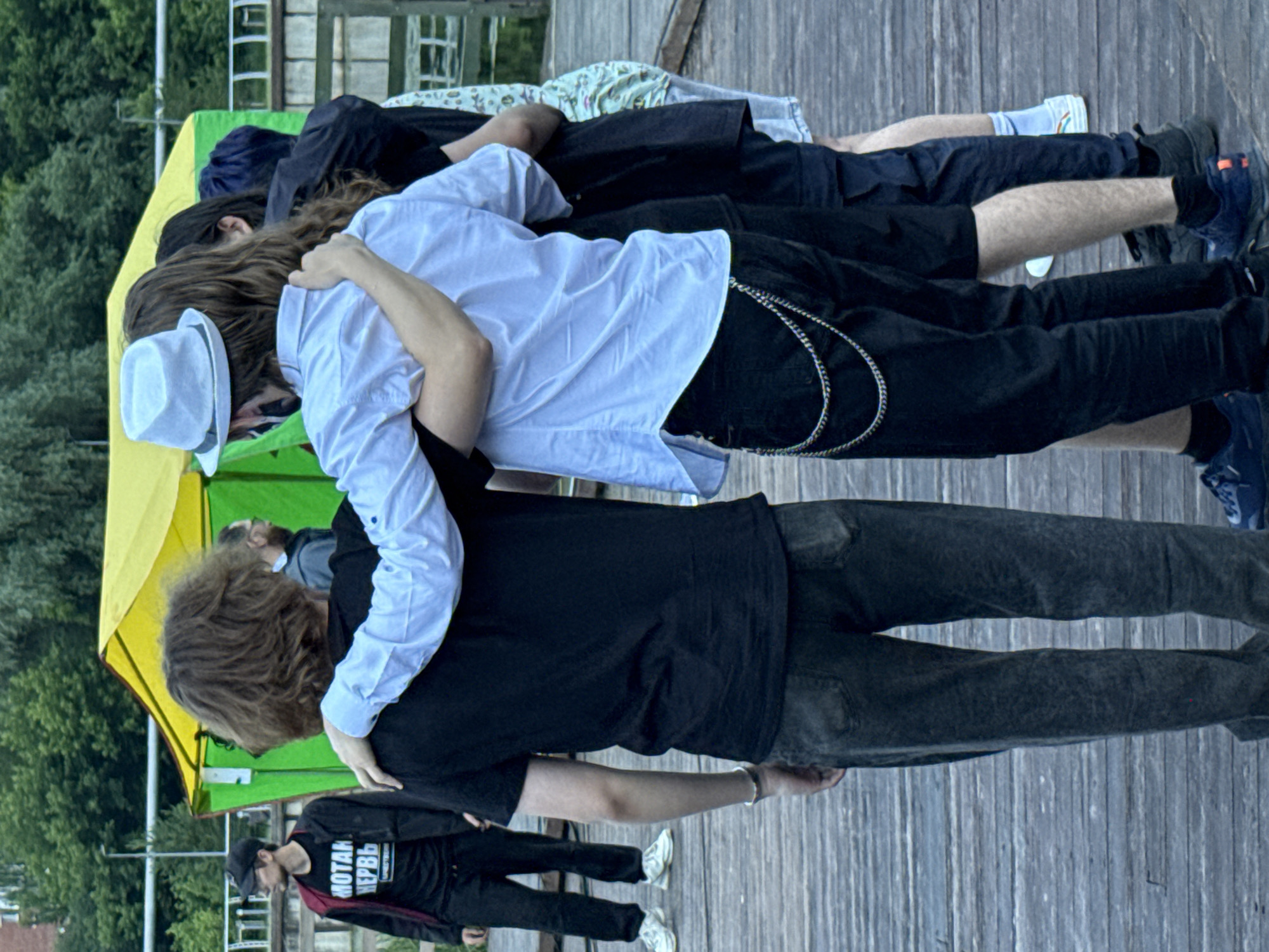The image depicts a group of five individuals standing on a wooden boardwalk. The individuals are primarily seen from the back, with their faces obscured or turned away.
In the foreground, two individuals are prominently featured, with one person's arm draped around the other's shoulders. The person on the left wears a black t-shirt and dark, possibly denim, pants. The person on the right wears a white collared shirt, black pants adorned with a silver chain, and a white fedora. Their postures suggest an embrace or close companionship.
Behind them, three other individuals are visible. One person, to the far left, wears a black baseball cap, glasses, and a black jacket over a black t-shirt with Russian text. They are looking to the left. To the right of this individual, another person is partially visible, wearing dark clothing. The furthest individual on the right wears a patterned shirt and light-colored shorts or skirt.
The environment appears to be outdoors, with a green and yellow tent structure visible in the background. Trees and foliage are also present, suggesting a park or natural setting. The boardwalk is made of weathered wooden planks.
The presence of Russian text on the t-shirt of the individual on the far left may suggest a connection to Russia, but a definitive identification of the location as Votkinsk, Russia cannot be made solely from the visual information provided. The scene appears to be a casual gathering of young people.