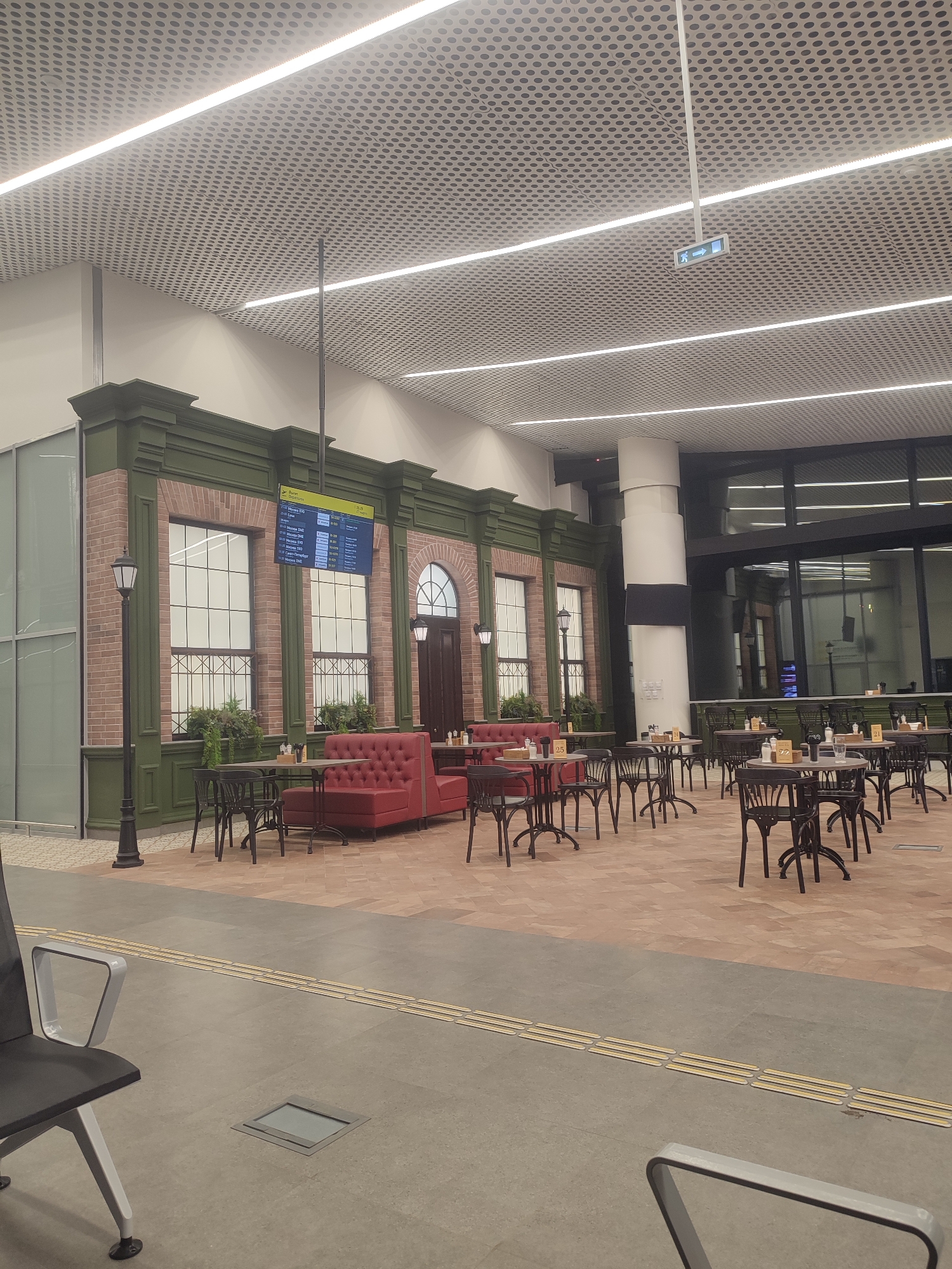 This image depicts the interior of a modern airport terminal, specifically the Izhevsk International Airport in Izhevsk, Russia. The scene is an unoccupied cafe or waiting area.
The environment features a high ceiling constructed from perforated white metal panels, illuminated by multiple parallel linear LED light fixtures. A green exit sign with a white arrow and person icon hangs from the ceiling in the upper right quadrant. The floor transitions from a light grey tile in the foreground, containing a tactile paving strip for the visually impaired and a square floor-level access panel, to a patterned reddish-brown tile in the mid-ground.
The central feature is a large, elaborate architectural facade designed to resemble a historical building. This facade displays sections of red brickwork interspersed with dark olive-green painted trim, paneling, and moldings. Three large, multi-paned window sections, apparently faux, are visible, with planter boxes containing artificial greenery positioned beneath them. A digital flight information display (FIDS) screen is mounted above the central window, showing "Вылет Departures" and listing several flights to Moscow airports (SVO, DME, VKO), including their flight numbers and departure times. To the left of the facade, a tall, black decorative lamppost with a lantern head stands. An arched, dark brown door is situated on the right side of the facade, flanked by another wall-mounted lantern.
In front of this facade, numerous small dining tables with light-colored circular tops and dark, ornate metal bases are arranged. Each table is accompanied by multiple dark metal chairs with intricate backrests. Several red, tufted upholstered booth seats line the facade wall, paired with tables. Condiment sets (shakers, napkin dispensers), coasters, and sometimes glasses are present on most tables, some bearing table numbers such as "25," "77," and "21." No individuals are present in the seating area or visible elsewhere in the image, indicating the area is currently empty.
In the background to the right, a large white cylindrical column rises to the ceiling, featuring a black band wrapping around its lower section. Behind this column, a glass wall with dark frames overlooks another section of the terminal or a departure gate area. Reflections are visible in the glass panels, indicating interior lighting.