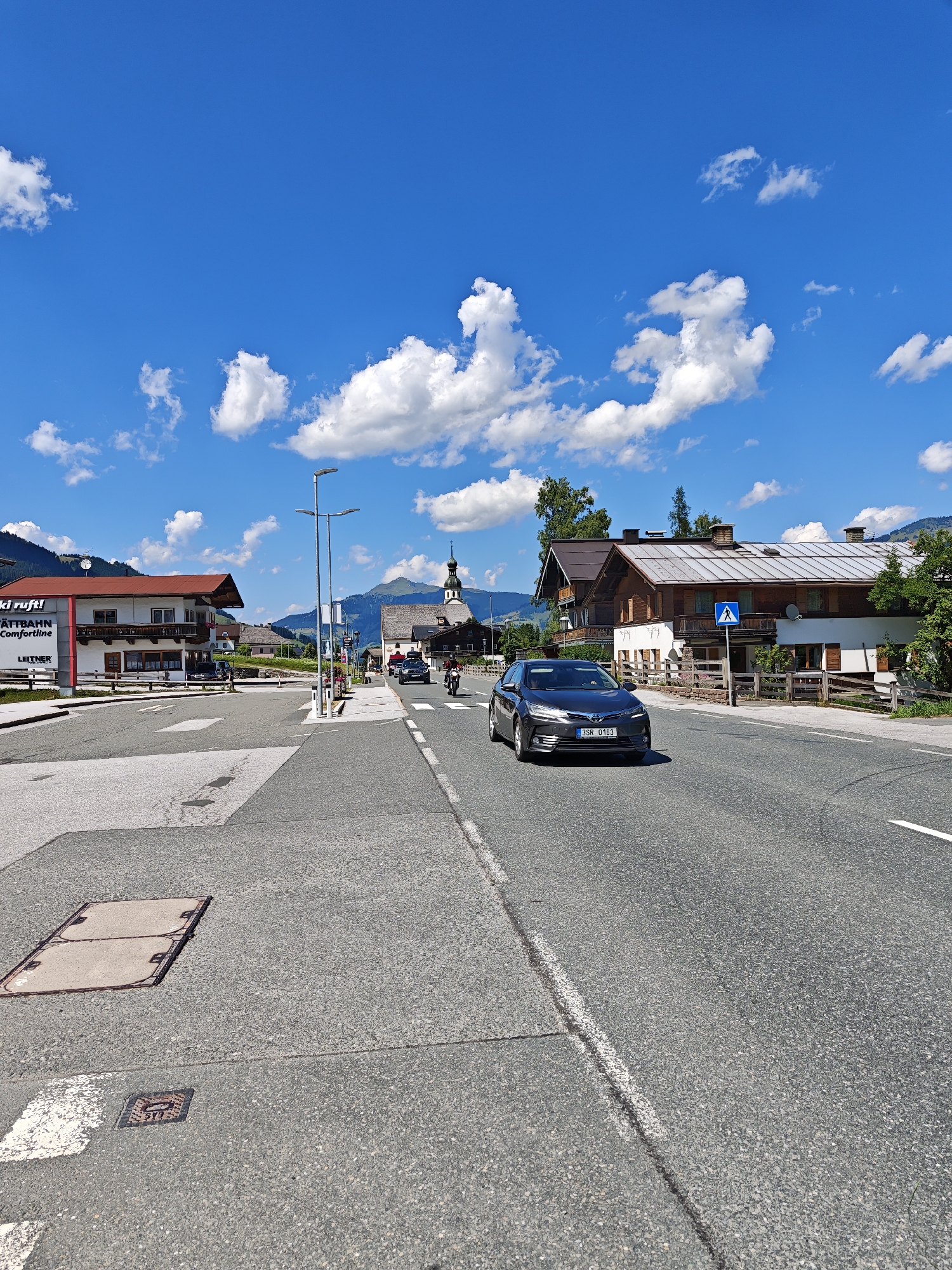 The image depicts a sunny day in Filzen, Austria, with a bright blue sky dotted with white clouds. A paved road runs through the scene, with a dark gray sedan driving on the right lane and a motorcycle on the left. On the left side of the road, there are buildings, including one with a red roof and a sign that reads "Ski ruft! GÄTTBAHN Comfortline LEITNER". Further down the road, a church with a steeple is visible, surrounded by traditional Austrian architecture. The landscape is mountainous, with green hills and trees in the background. The road has white lane markings, and there are streetlights and a pedestrian crossing sign. The ground on the left side of the road has manhole covers.