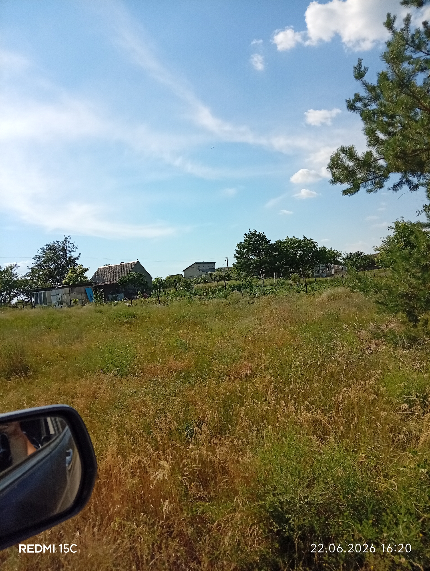 The image depicts a rural landscape with several buildings, overgrown vegetation, and a clear blue sky with scattered clouds. In the foreground, the side mirror of a vehicle is visible, reflecting a person's arm. The scene appears to be a sunny day, likely in the late afternoon given the timestamp.
The subjects include a few rustic buildings, one with a grey, sloped roof, and another further in the background that is more modern. The vegetation is a mix of tall, dry grasses and green bushes, suggesting a natural, unmanicured setting. There are also several trees, some appearing to be fruit trees or deciduous, and a prominent pine tree on the right side of the frame. A wire fence can be seen running through the mid-ground.
No specific activity or event is overtly taking place; the scene suggests a quiet, everyday moment in a rural environment. The overall mood is peaceful and serene. The time of day is indicated by the timestamp "22.06.2026 16:20," suggesting it is late afternoon. The weather appears to be fair and sunny, with a bright sky and fluffy white clouds. The location context is Mykolaivka, Ukraine, which aligns with the rural aesthetic. Visible text includes "REDMI 15C" in the bottom left corner and the date and time in the bottom right.