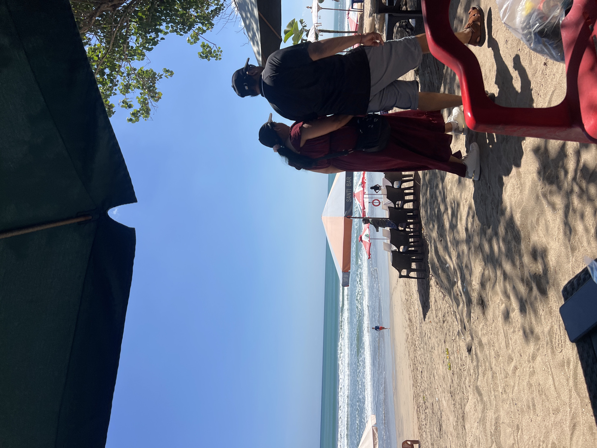 In Kuta, Indonesia, a sandy beach is captured under a clear blue sky, with the turquoise ocean meeting the shore in gentle waves. Two people, a man and a woman, walk away from the camera, their backs to the viewer. The woman wears a long, maroon dress and a dark baseball cap, while the man sports a black t-shirt, a black baseball cap, and light grey shorts. They are walking on the sand, with the ocean and beach umbrellas visible in the background. Several beach chairs and tables are set up on the sand, suggesting a cafe or restaurant setting. Further down the beach, a lone figure in a red and blue outfit runs towards the water. The foreground is dominated by a red plastic chair and a table, partially obscuring a portion of the scene. Shadows from palm trees are cast across the sand, adding depth to the image.