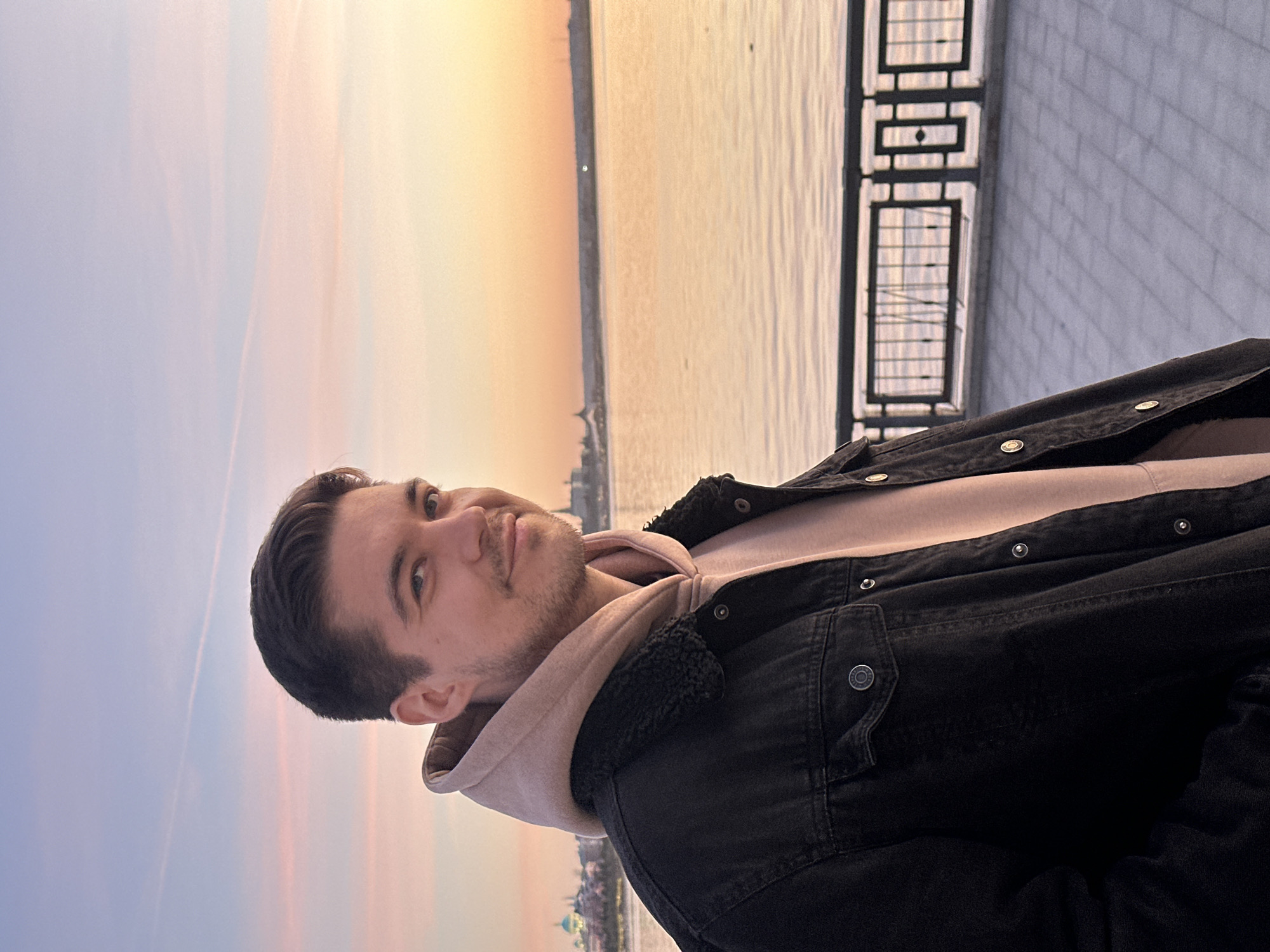 The image, oriented vertically, captures a young man posing on an urban embankment during sunset or sunrise. The sky dominates the upper portion, displaying a vibrant gradient from soft purple and blue on the left to warm orange and yellow on the right, indicative of the golden hour. Faint, wispy cloud trails are visible in the sky.
The main subject, a man in his late teens or early twenties, is positioned on the left side of the frame. He has short, dark hair and light stubble/a short beard. He is wearing a black denim jacket with a dark, possibly sherpa-lined collar, over a light pink or beige hooded sweatshirt. He looks directly at the camera with a slight, calm smile, his expression relaxed and content.
Behind the man and extending across the right half of the image is a wide body of water, likely a river or large lake, reflecting the warm hues of the sky. The water appears calm with subtle ripples. On the far bank, a distant cityscape is visible, featuring several buildings with distinct architectural elements, including what appear to be domes and spires, suggesting significant historical or religious structures. Given the context, these could be landmarks in Kazan, Russia, such as those within the Kazan Kremlin. A dark metal railing or fence separates the paved embankment from the water on the far right of the image. The pavement appears to be made of light-colored bricks or cobblestones.
The overall scene conveys a peaceful and contemplative mood, typical of an evening or morning stroll by the water in a city setting. No visible text is present in the image.