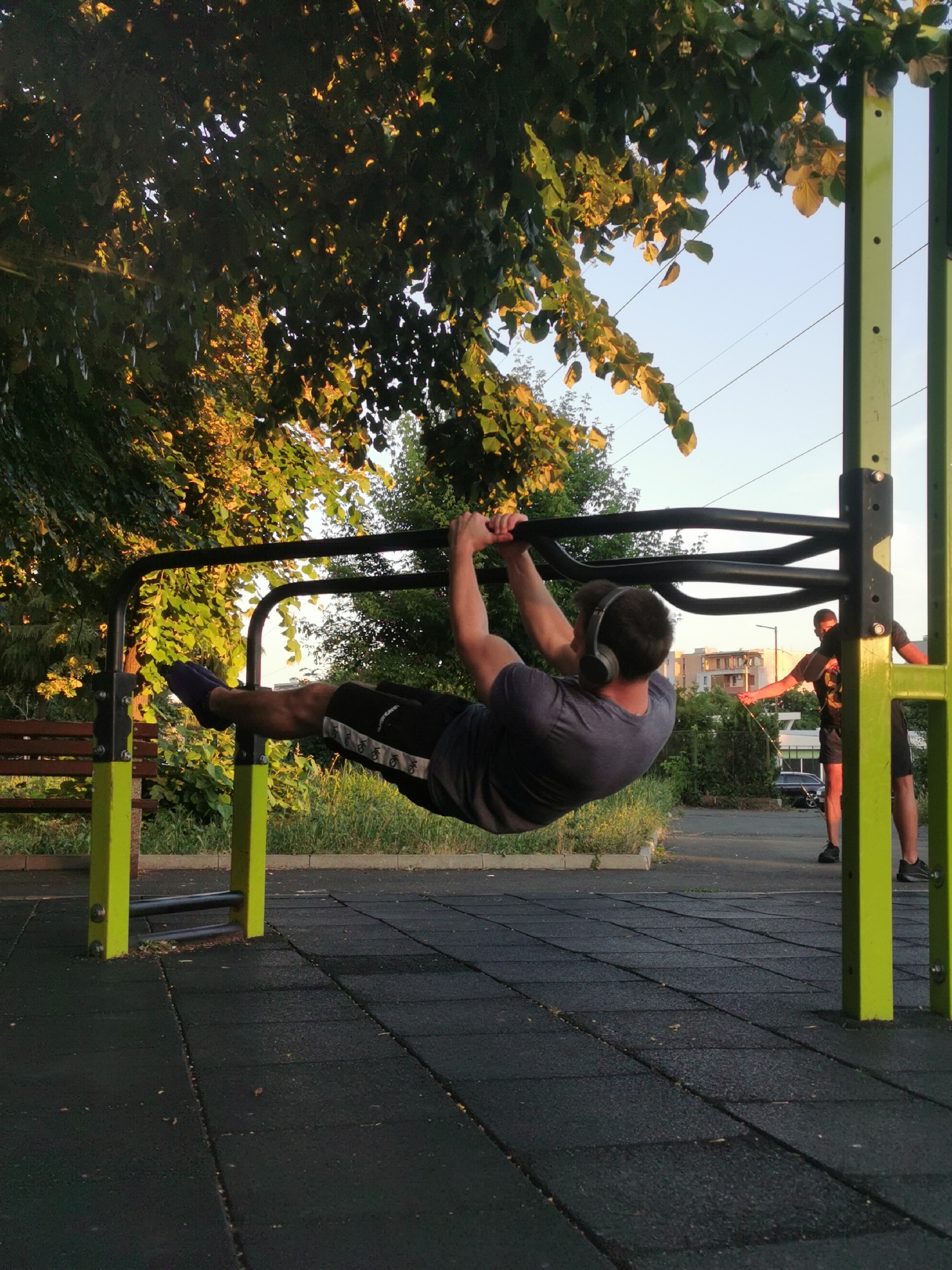 A man wearing headphones is performing an L-sit exercise on a pull-up bar at an outdoor gym. He is wearing a dark t-shirt and black shorts with a white stripe. His legs are extended straight out in front of him, and his body is parallel to the ground. Another man is standing in the background to the right, appearing to interact with someone off-camera. The gym equipment has bright yellow support posts and black bars. The ground is covered with black rubber matting. Trees with green and yellowing leaves are visible behind the pull-up bar, and in the background, there are buildings and a clear sky. The setting appears to be a park or public outdoor fitness area. This scene was captured in Burgas, Bulgaria.