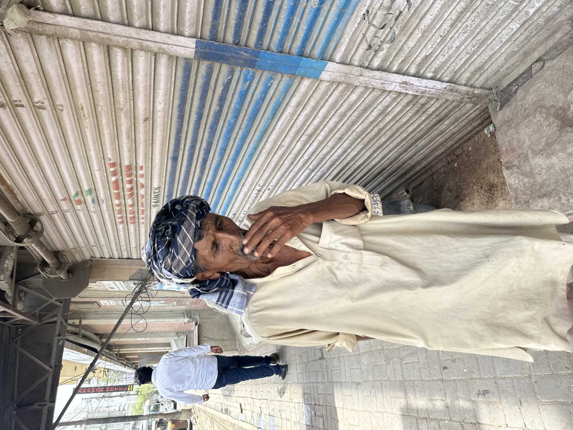 An outdoor, medium shot captures two men walking on a paved sidewalk in front of a closed corrugated metal storefront. The man in the foreground, an elderly South Asian male with a turban, faces the right side of the frame with his hand near his mouth. He is wearing a light-colored, knee-length tunic over loose-fitting pants and a patterned scarf that is part of his turban. The man in the background, dressed in a white collared shirt and dark pants, walks away from the camera toward the left side of the frame. The storefront features blue and white stripes and is marked with faded red lettering that includes the word "PAINTS." The environment appears to be an urban street scene in Karachi Division, Pakistan, with a visible overhead structure and electrical wires suggesting a busy commercial area.