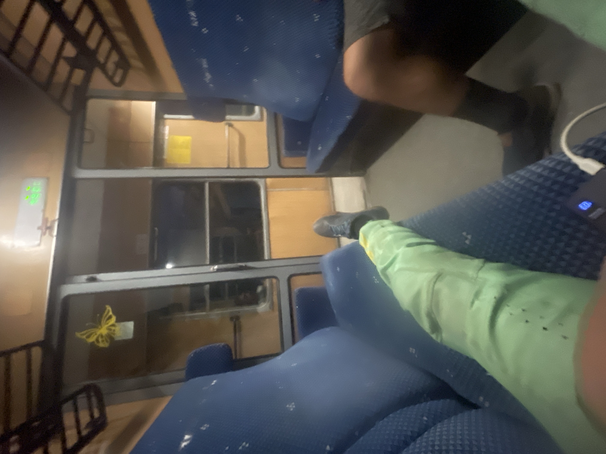 An interior view of a train carriage shows blue upholstered seats and overhead luggage racks. A yellow butterfly sticker is visible on a glass partition. The lighting suggests it is evening or nighttime. In the foreground, a person's legs are visible, with one leg clad in a light green garment and the other in dark socks and shoes. A black electronic device with a white cable is also in the foreground. The train carriage appears to be stationary. The image does not provide any specific information about the city of Borovo, Bulgaria.