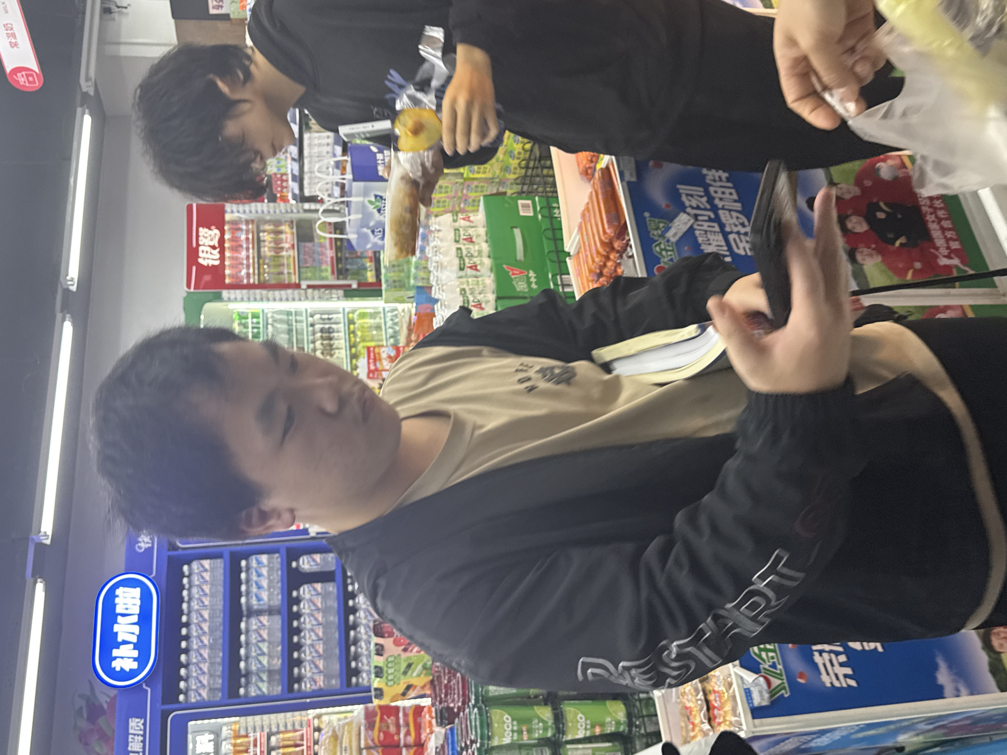A medium shot, taken at eye-level, shows a man holding a smartphone while in the aisles of a Chinese convenience store. The man is wearing a black windbreaker over a tan t-shirt. In the background, another person wearing a black shirt holds food. Shelves containing beverages and various other food items are visible. The store is in Jiangjin, China. The shelves have signs in Chinese.