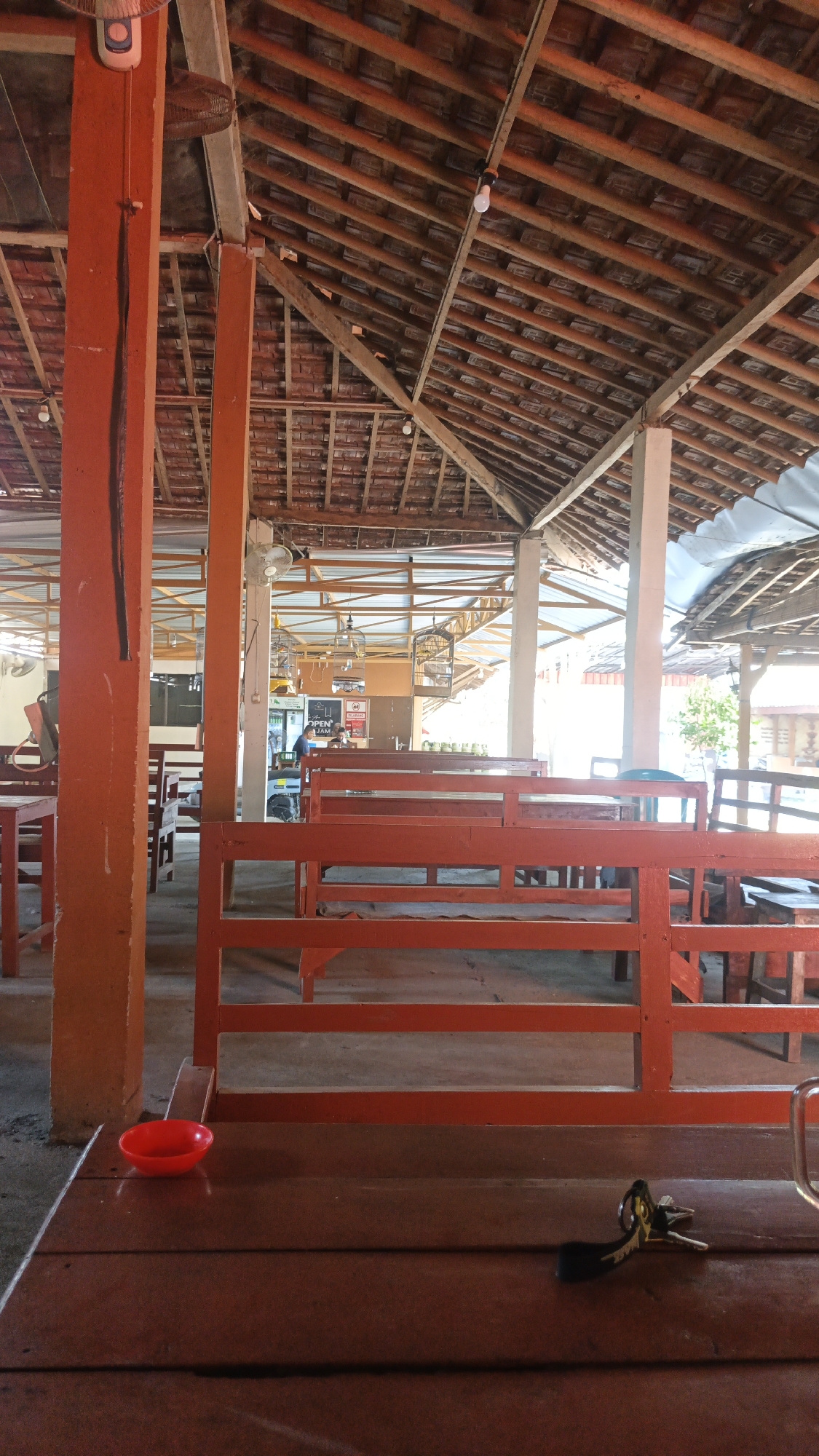 The image depicts the interior of an open-air food stall or restaurant in Ngembalrejo, Indonesia. The structure features a prominent reddish-orange wooden pillar in the foreground, supporting a high, tiled roof supported by exposed wooden beams and rafters. Rows of similar reddish-orange wooden benches and tables are visible, arranged in an organized manner. In the background, more seating is apparent, along with some caged birds hanging from the ceiling and a man standing near a counter. A small red bowl rests on one of the tables in the foreground, and a set of keys lies on the wooden surface near the bottom right of the frame. The overall impression is that of a simple, functional, and somewhat rustic establishment, typical of many local eateries in Indonesian towns.