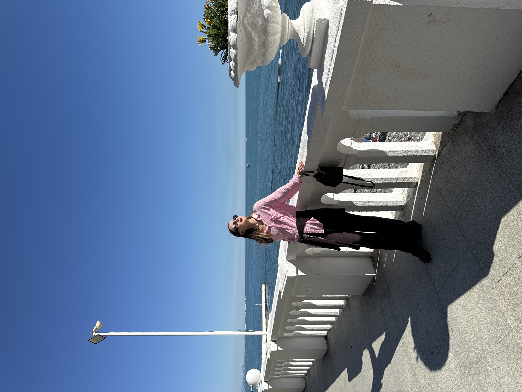 The image, when properly oriented, depicts a young woman posing on a sunny seaside promenade in Sochi, Russia. She is positioned with her back arched slightly, leaning against a white, classical-style balustrade, and gazing upwards towards the clear blue sky. She is dressed in a pink long-sleeved button-up shirt, dark trousers, and dark shoes, with sunglasses on her face. A dark handbag hangs from her left shoulder, resting on the balustrade.
The setting is a vibrant outdoor scene overlooking the sea. The white balustrade features decorative columns and a sturdy top rail, extending along the foreground. Beyond the balustrade, the deep blue, calm sea stretches to the horizon, dotted with a few small boats in the distance. A pebble beach is partially visible below the balustrade. To the right, a large white stone planter brimming with yellow flowers and green foliage is placed on the balustrade. On the left, a tall white lamppost equipped with a light fixture and a horn-like speaker extends into the cloudless sky.
The scene is brightly lit, indicative of midday or early afternoon, with strong, distinct shadows cast by the woman and the balustrade onto the grey, textured paved ground. The weather is clear and sunny, contributing to a cheerful and relaxed atmosphere. No visible text is present in the media file.