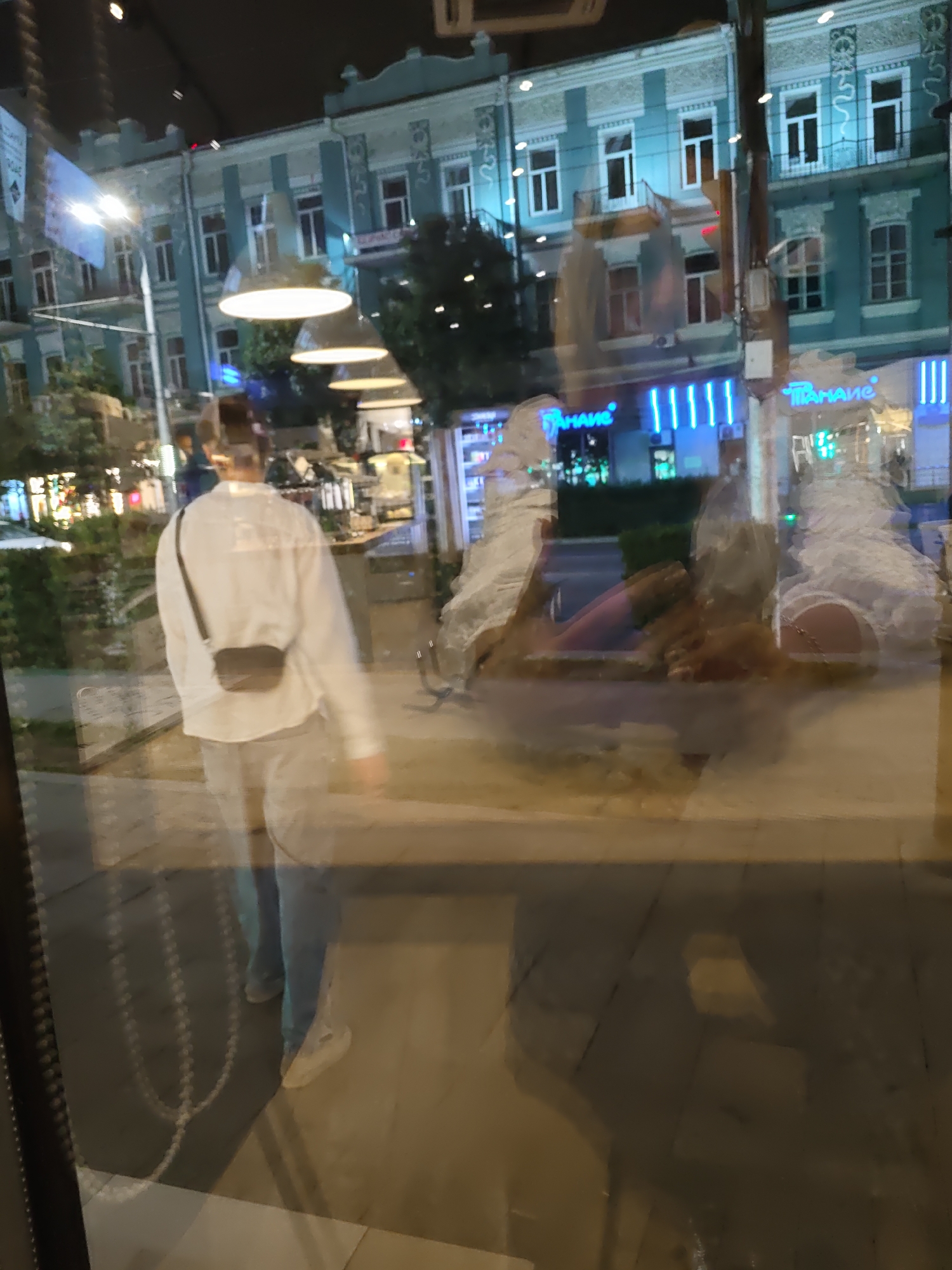 The image captures a street scene at night in Rostov-on-Don, Russia, as seen through a reflective surface, likely a storefront window. The reflection shows a person wearing a white long-sleeved shirt and light-colored pants, with a small dark bag slung across their body. The person is walking away from the camera. Behind them, the reflection reveals a building with teal facades, white window frames, and decorative elements. Neon lights, with Cyrillic script reading "Таимаиг," are visible on the ground floor, suggesting commercial establishments. The street is paved, and trees are visible in the background, illuminated by streetlights. The overall impression is that of a well-lit urban environment at night.