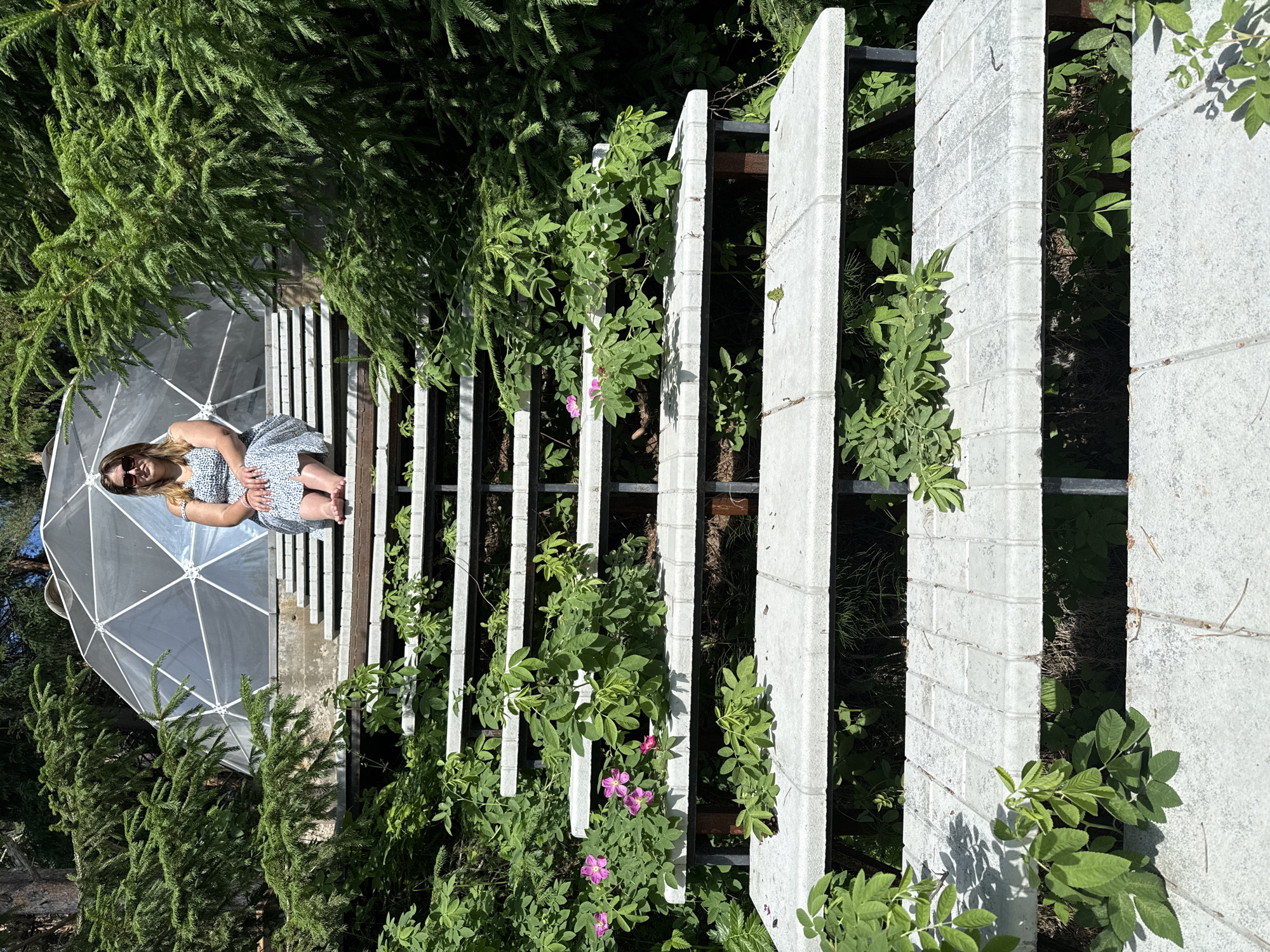 A woman with blonde hair and sunglasses is sitting on a set of concrete stairs. She is wearing a blue and white patterned dress. Behind her, a geodesic dome structure is visible. The stairs are surrounded by lush green foliage and trees. The scene appears to be outdoors. There is no information available to determine the location in Makarovo, Russia.