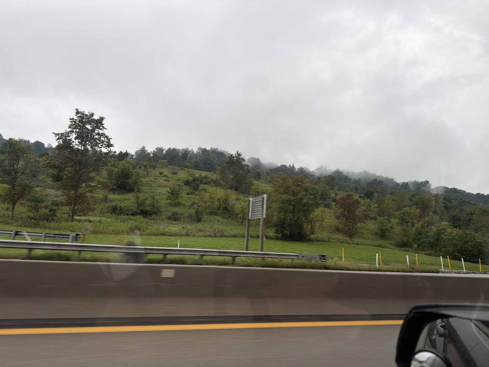 The image captures a view from a vehicle moving on a highway. In the foreground, a concrete barrier separates the highway lanes from the shoulder. A yellow line marks the edge of the traffic lane. In the middle ground, a guardrail runs along the side of the highway, with a grassy embankment behind it. The embankment is dotted with trees and shrubs, and further up the hill, the landscape is covered in dense foliage. The sky is overcast and appears misty or foggy, especially in the distance where the trees on the hills blend into the atmospheric haze. The scene is a natural landscape along a highway, characteristic of a rural or semi-rural environment. The location is Fallowfield Township, United States of America.