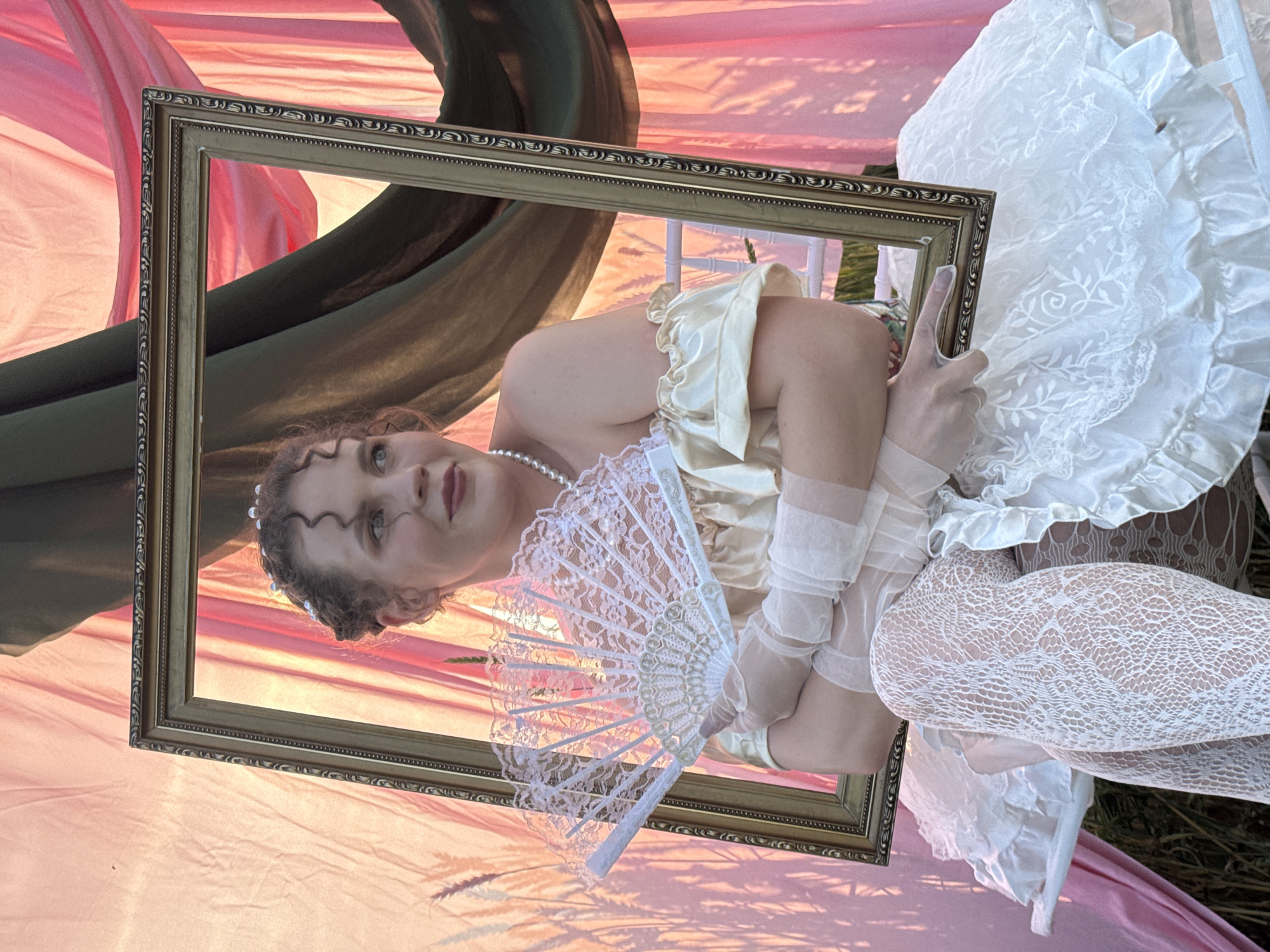 A young Caucasian woman with curly auburn hair and fair skin is pictured holding a white lace fan and a gold-colored frame. She is wearing a yellow, off-the-shoulder dress with ruffles, sheer white opera gloves, and white lace stockings. Her hair is styled up with small pearls. She is seated on a white bench with ruffles, and the background consists of draped pink and dark green fabric. The lighting suggests it is either dawn or dusk. There is no identifiable indication of the location within the city of Foki, Russia.