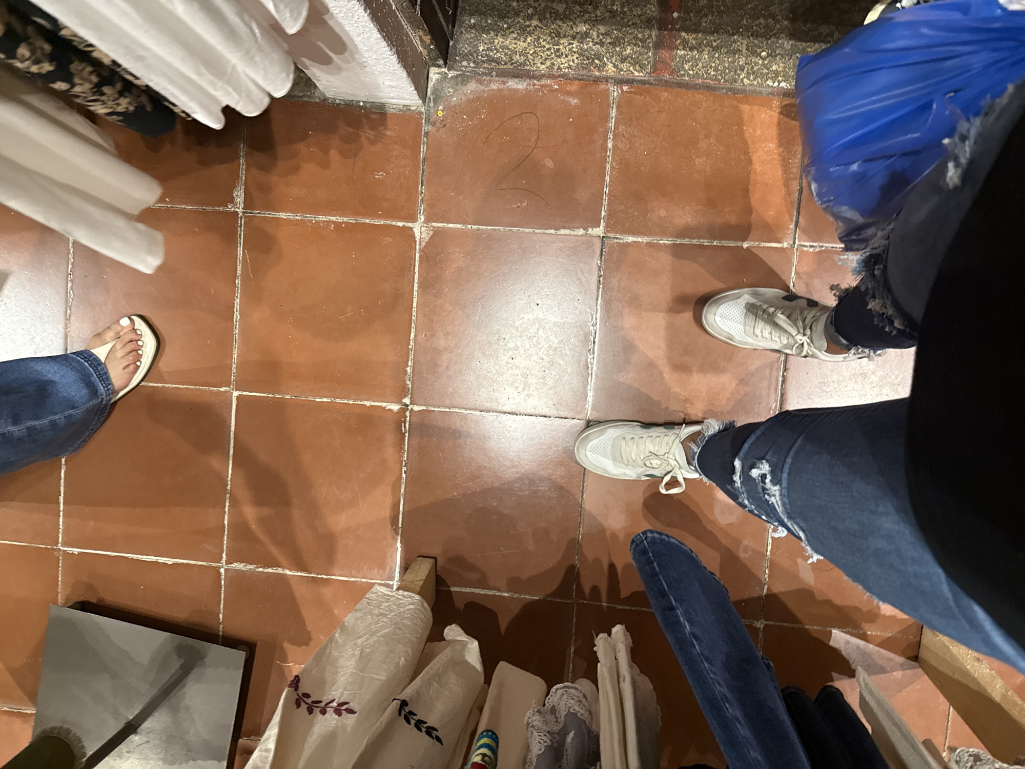 The image shows an indoor scene of a retail space with terracotta tile flooring. The composition is a high-angle shot capturing the lower portions of three individuals and various objects on the floor. One person is wearing blue jeans and a sandal with white toenail polish. A second individual is wearing distressed denim jeans and white sneakers. A blue plastic bag sits near this person. The third person is wearing dark-colored jeans and white sneakers. Rolls of fabric are scattered on the floor, some appearing to be on stands. The scene's location is inferred to be a retail shop or market in San Cristóbal, Mexico, although there are no elements of the image to directly confirm this.