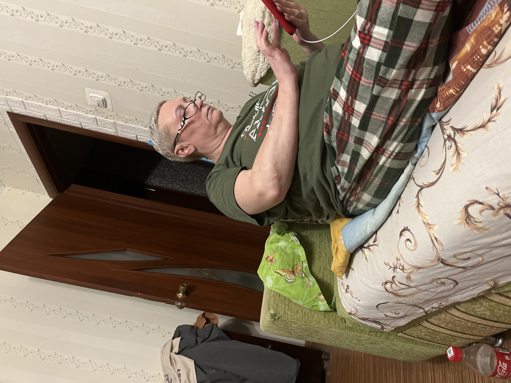 A woman with short blonde hair and glasses is sitting on a couch and looking at a red tablet. She is wearing a green t-shirt and plaid pants. The couch is covered with a floral patterned blanket and a green cloth with butterflies on it. There is a bottle of Coca-Cola on the floor next to the couch. The room has striped wallpaper and a wooden door. The image appears to have been taken indoors, possibly in a residential setting in Minsk, Belarus.