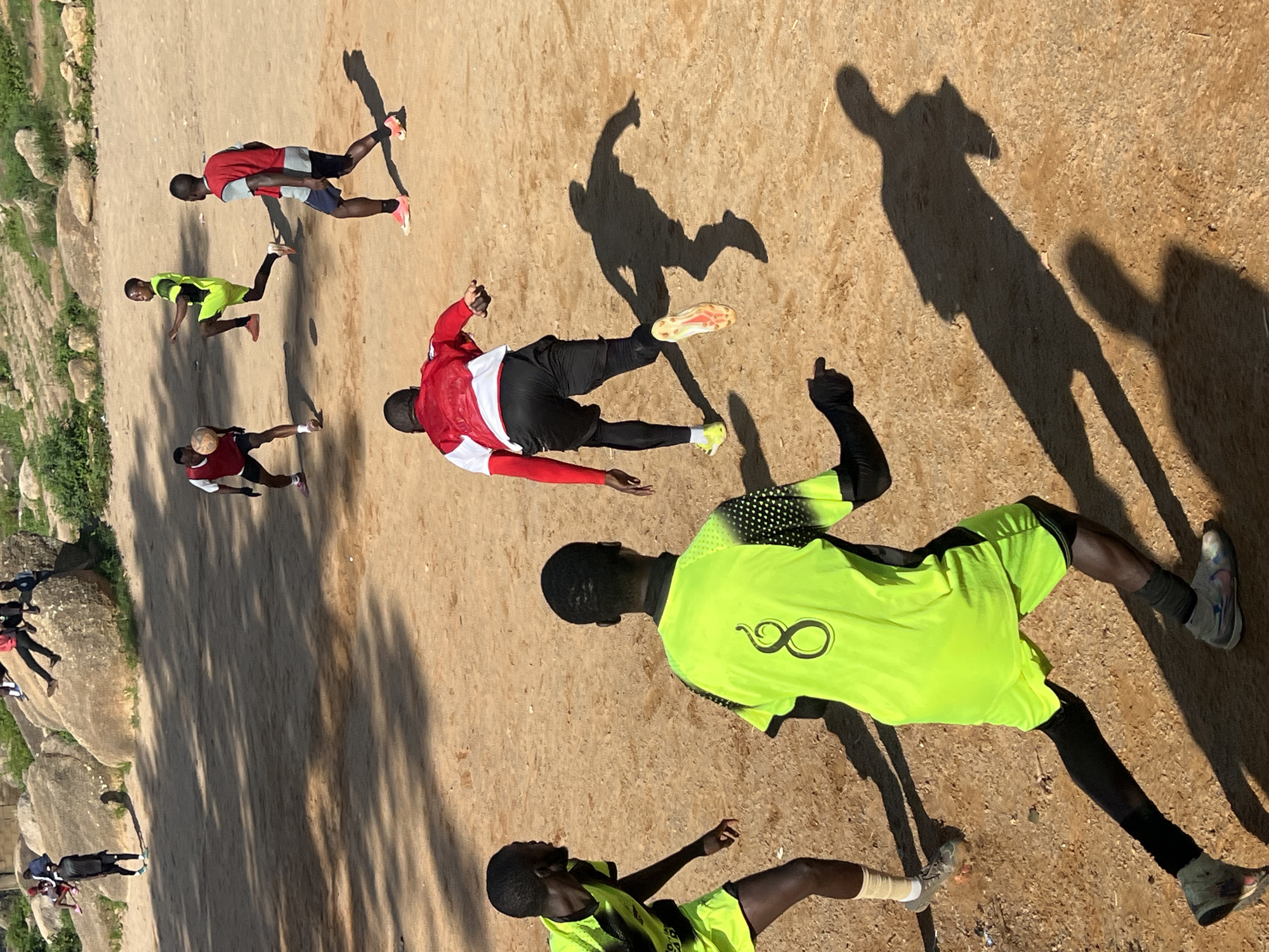 The image depicts a group of young men playing soccer on a dirt field in Jos, Nigeria. The field is uneven and appears to be a natural clearing surrounded by rocky terrain and sparse vegetation. The players are dressed in athletic wear, with some wearing vibrant jerseys and shorts, while others are in simpler attire. The sun is casting long shadows, indicating it is likely late afternoon. The overall atmosphere appears to be one of casual recreation, with the players actively engaged in the game. The terrain suggests an informal setting, possibly a community field or a less developed sports area within Jos.