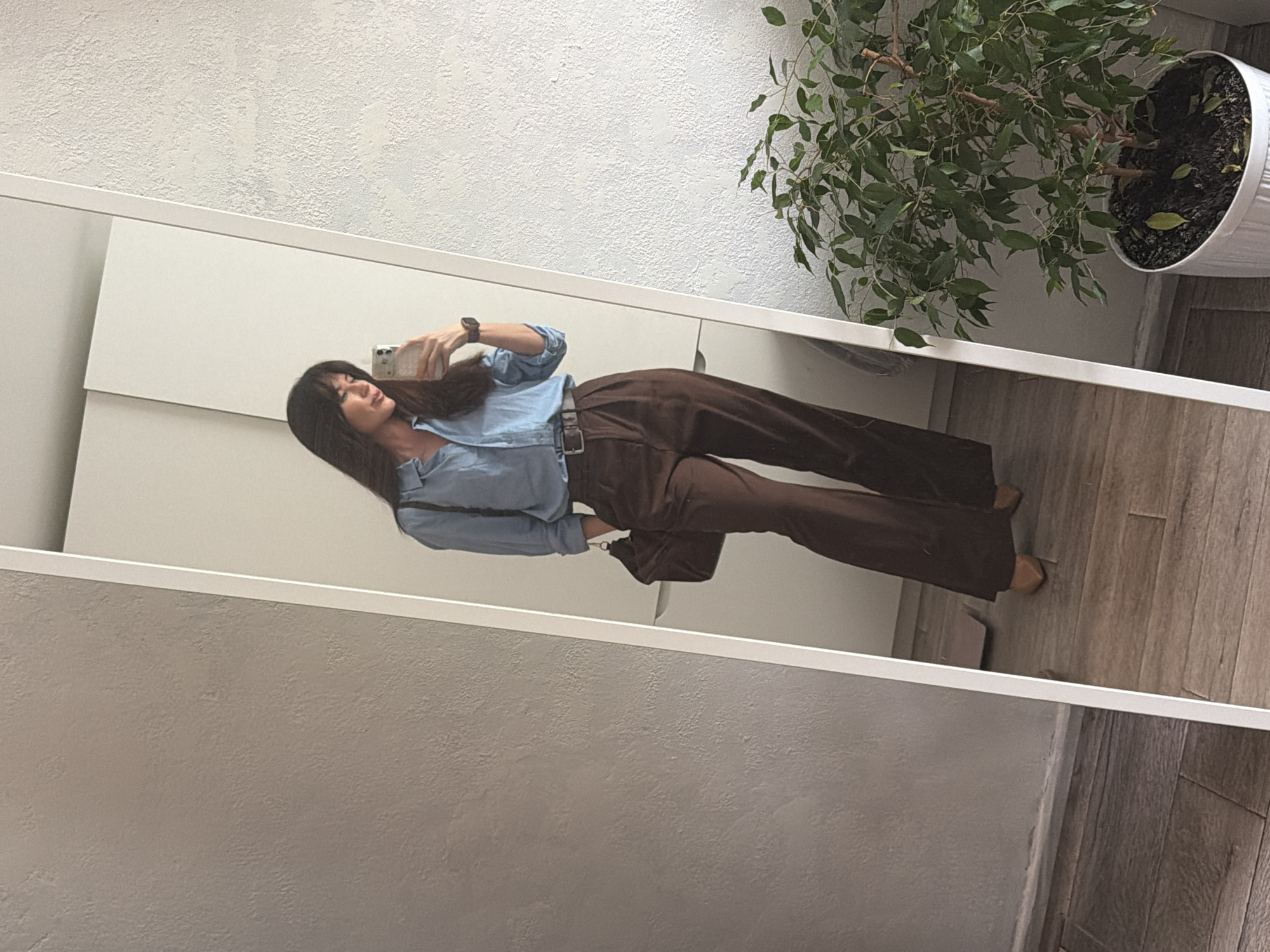 A woman is reflected in a mirror. She is holding a smartphone and wearing a light blue shirt, brown trousers, and a watch. A potted plant is visible in the upper right corner. The location is Perm, Russia. The background walls are textured and painted white. The floor appears to be wood-like.