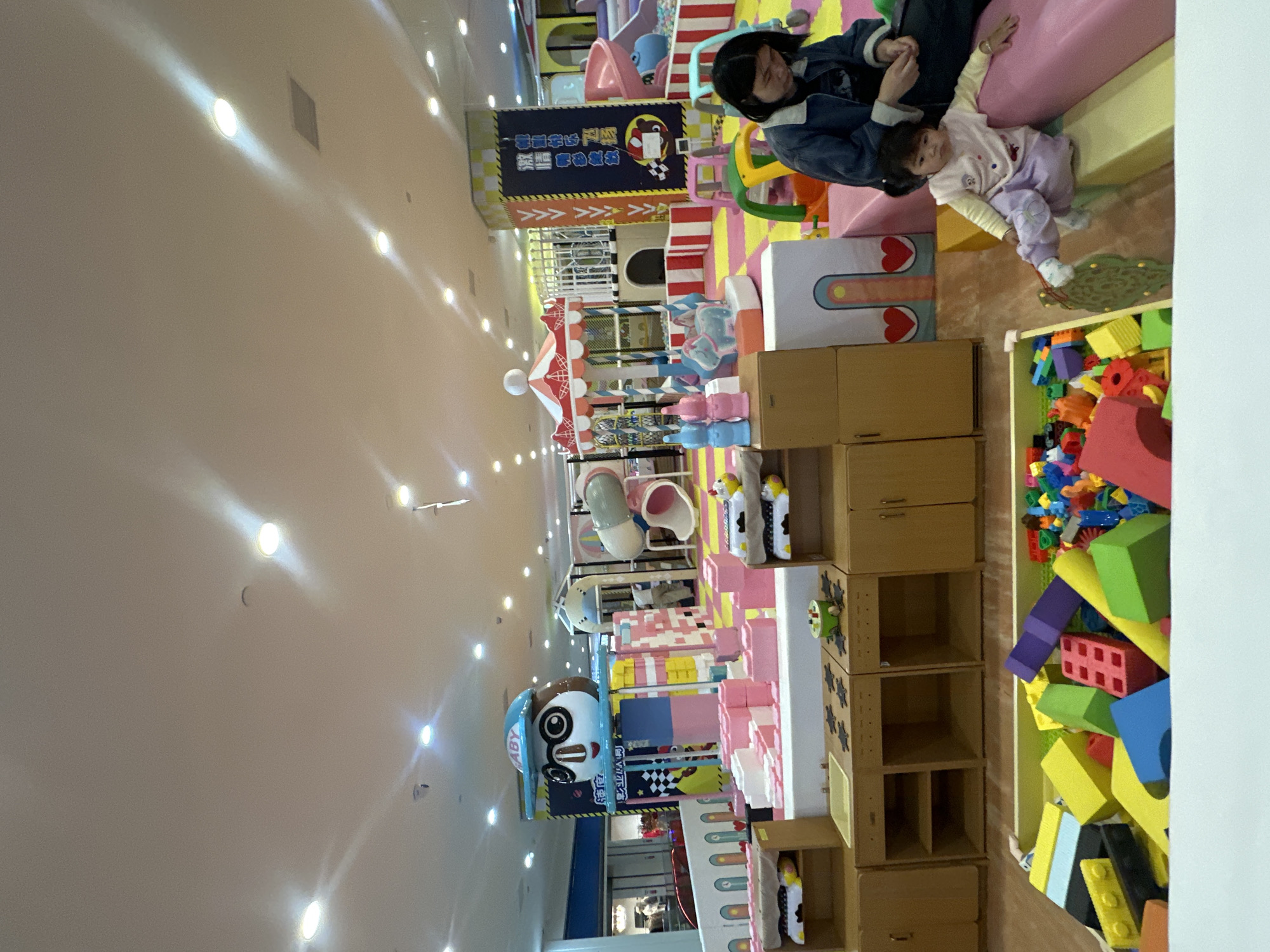 The image depicts an indoor children's play area, likely located within a commercial establishment in the Chongqing High-tech Zone, China. The scene is characterized by a vibrant and colorful environment filled with various play structures and toys. In the foreground, a toddler is seated on a pink cushioned bench, looking down. To the left of the toddler, a large bin overflows with an assortment of colorful building blocks, including large Lego-style bricks and smaller plastic pieces. Behind the toddler, an adult, presumably a parent or guardian, is seated on the same bench, looking down towards the child.
The play area features a carousel with red and white stripes, a small blue horse sculpture, and several pink and blue rabbit-shaped figures. Further in the background, a large cartoon character head with glasses and a blue cap stands atop a colorful play structure made of large building blocks in pink, white, and yellow. A tubular slide system in light gray and pink is also visible, leading into a ball pit filled with purple balls. The walls of the play area are decorated with colorful checkered patterns and cartoon graphics. Overhead, fluorescent lighting illuminates the space. The overall impression is one of a well-equipped and engaging environment for young children's recreation.