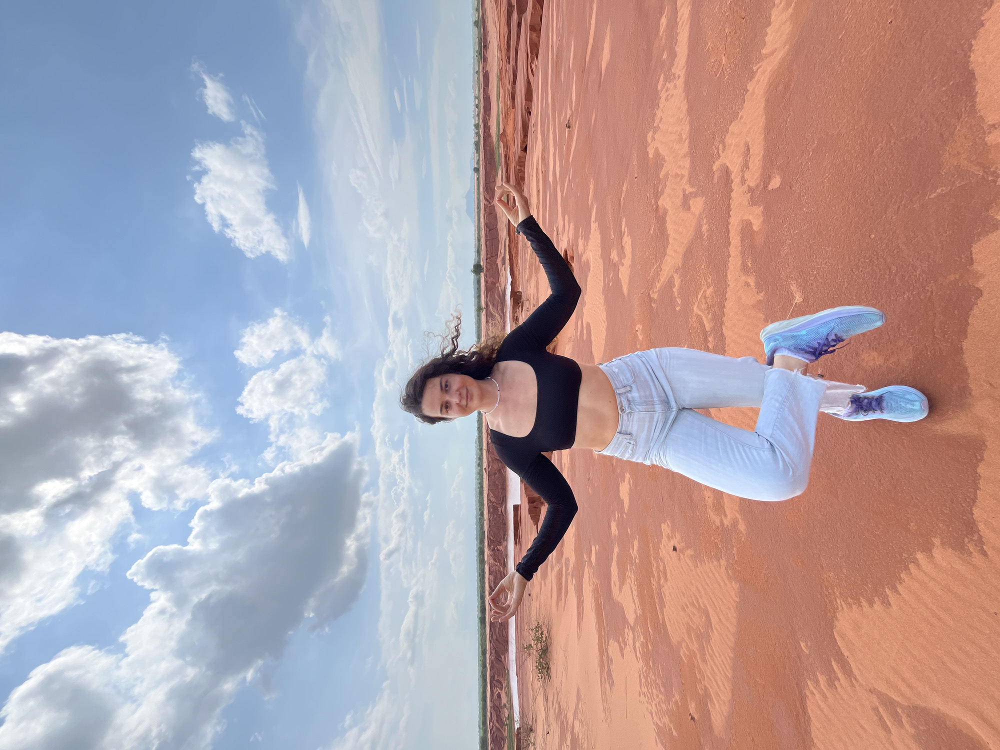 A young woman with long, dark, curly hair stands in a yoga pose on a sandy, reddish landscape under a bright blue sky with scattered white clouds. She is wearing a black long-sleeved crop top, light blue flared jeans, and colorful blue and purple athletic shoes. Her arms are extended to her sides with her palms facing upwards, and her right leg is bent at the knee, with her foot resting against her left calf. Her gaze is directed towards the camera with a slight smile. The background reveals a vast, barren terrain with undulating sand dunes and a body of water in the distance, characteristic of the sand dunes in Mui Ne, Vietnam. The lighting suggests it is daytime, with the sun casting subtle shadows on the sand.