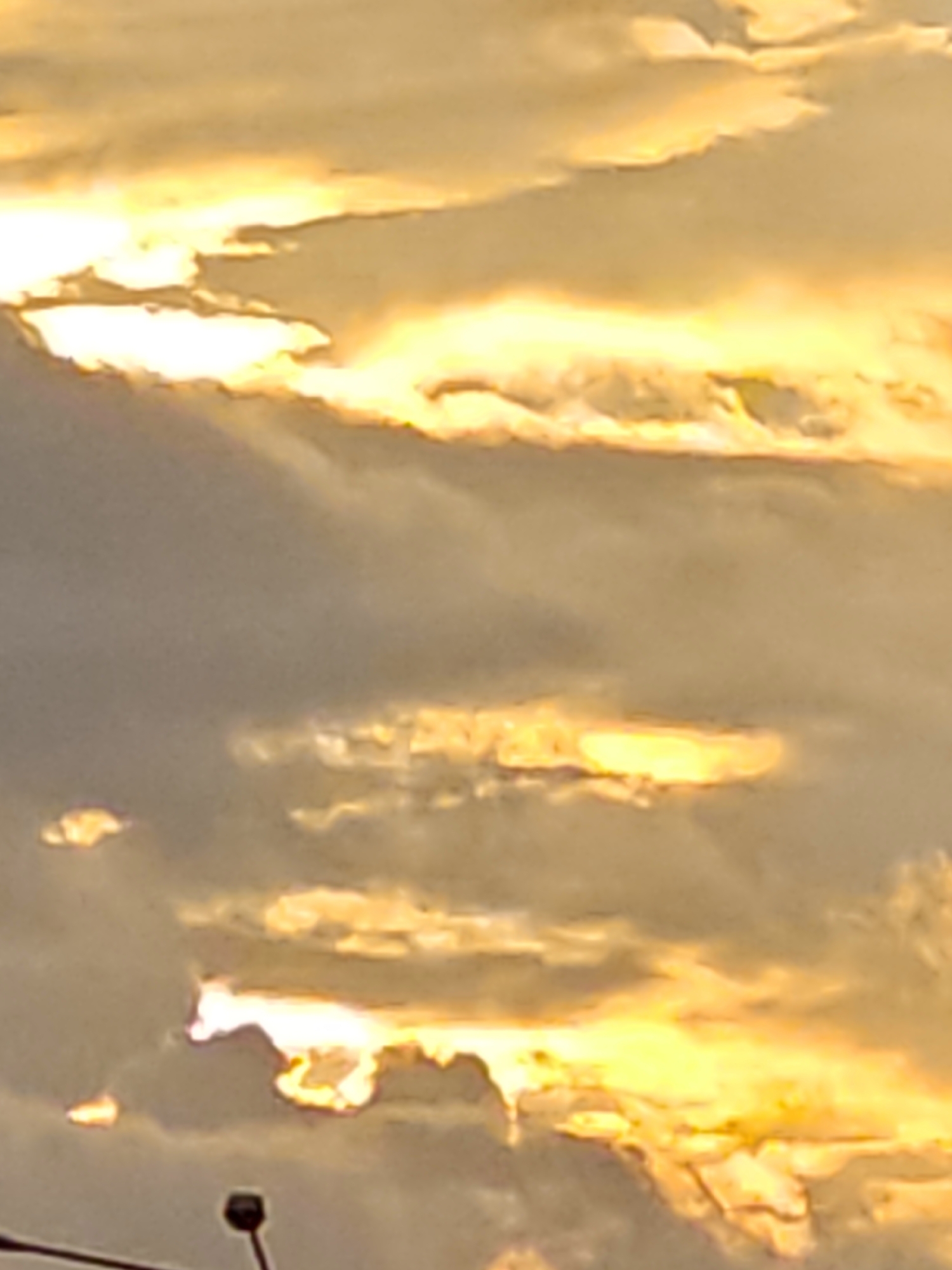 The image shows a cloudy sky with bright sunlight breaking through the clouds, creating streaks of golden and white light. The sky is a mix of gray and yellow tones, with some darker clouds interspersed. In the lower part of the image, a silhouette of a streetlamp or similar structure is visible, suggesting an outdoor urban setting. The overall impression is of a dramatic sky, possibly during sunrise or sunset. The image was taken in the city of Shcherbinka, Russia.