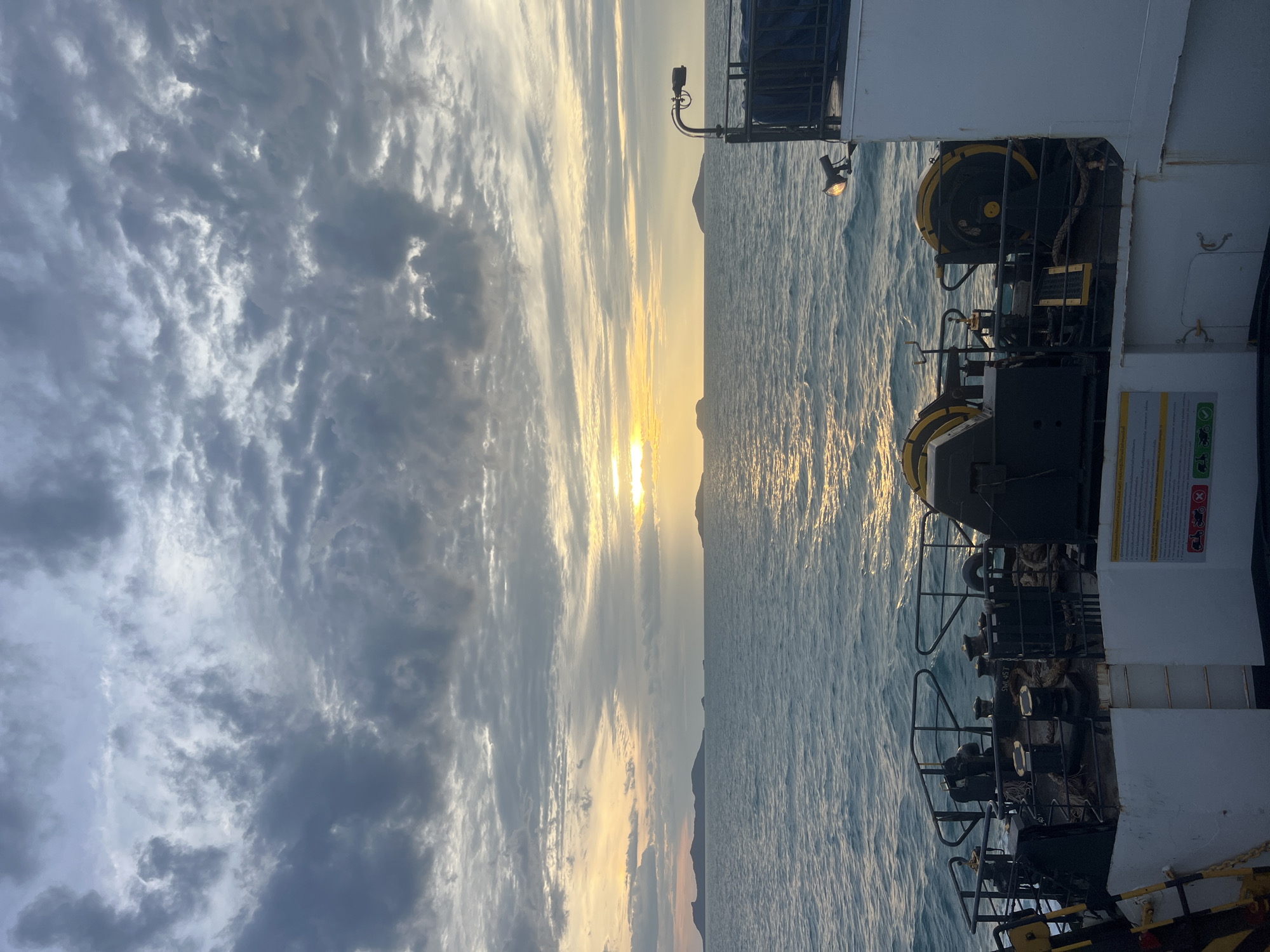 A wide shot captures a body of water and a cloudy sky at dusk. The perspective is from the deck of a ferry, looking out over the ocean. Several islands are visible in the distance, silhouetted against the orange and yellow hues of the setting sun. The water is choppy, reflecting the sky's colors. On the ferry's deck, various pieces of equipment are visible, including winches and ropes. The sky is filled with dramatic, dark clouds, with lighter patches where the sun is breaking through. The overall mood is serene and atmospheric.