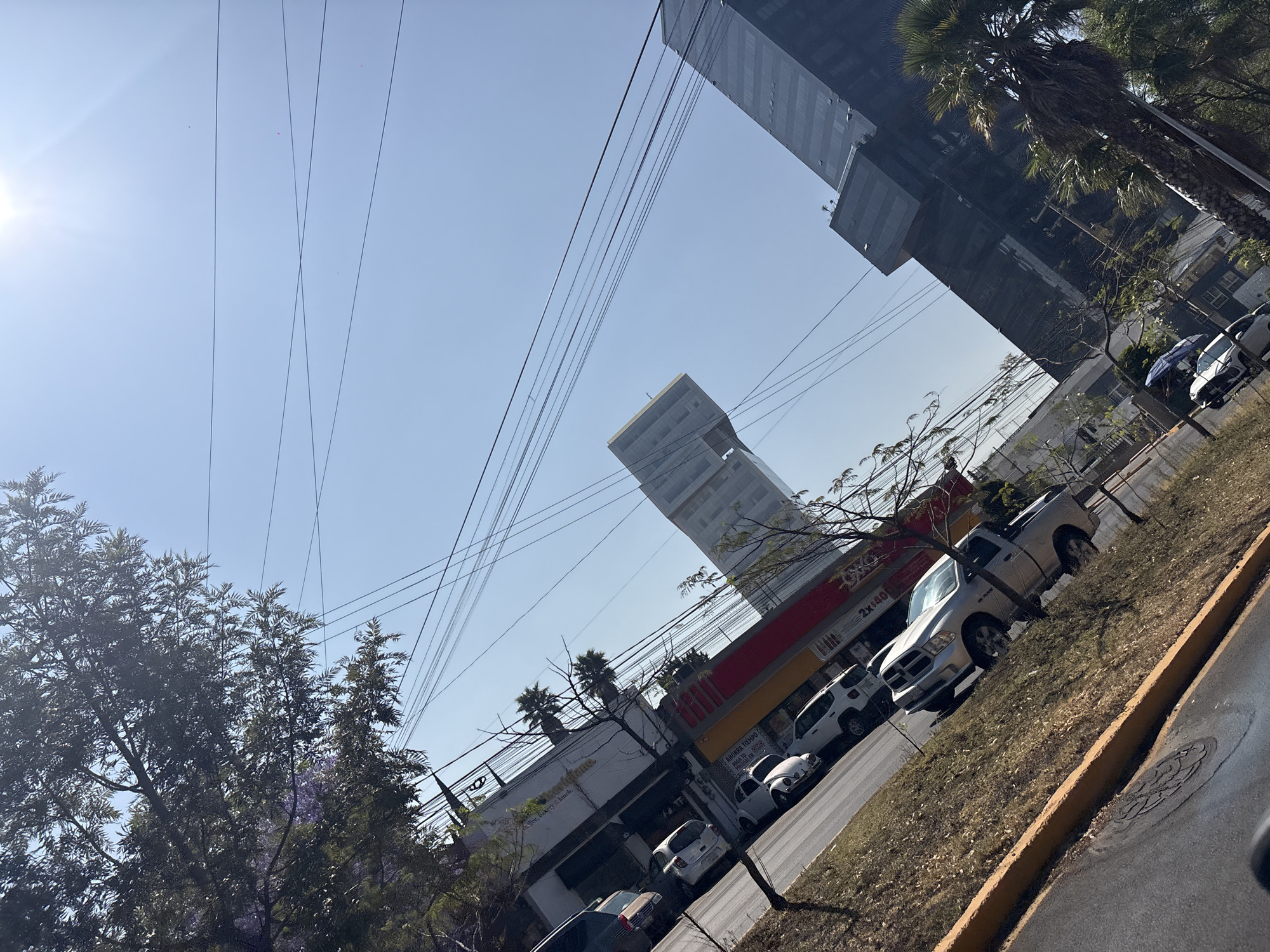 The image depicts an urban street scene in Puebla, Mexico, viewed diagonally upwards under a clear, bright blue sky with direct sunlight in the upper right.
In the background, two prominent modern high-rise buildings dominate the skyline. The building on the left is a lighter-colored structure with distinctive architectural elements at its peak. The building on the right is a dark, reflective skyscraper with a prominent, angular cantilevered section midway up its facade. These structures are consistent with the Torres JV in the Angelópolis district of Puebla.
Below the skyscrapers, a commercial strip runs parallel to the street. This low-rise building features a white upper section and a red and yellow lower facade. Visible signage includes "PASTELERÍA" (Bakery) and "PANADERÍA" (Bread Shop), along with "VENTAS Y ENCARGOS" (Sales and Orders) and a "2x$40" promotion on a red background.
A paved asphalt road segment is visible in the lower left foreground, featuring a circular metal manhole cover. A raised, grassy median or shoulder, with dry brown and sparse green vegetation, separates the road from the commercial buildings. A yellow-painted curb defines the edge of this median.
Several vehicles are parked along the street or shoulder. From left to right: a dark-colored sedan (partially obscured), a white compact car, an older model white Volkswagen Beetle, a white SUV, and a silver Dodge Ram pickup truck. Further down the street, a white car is parked, with a blue and white umbrella positioned near it.
Numerous black overhead power and communication lines crisscross the sky, extending between visible utility poles that are partially obscured by trees. Dark green foliage from various trees frames the upper left portion of the image, and a palm tree is visible on the right, in front of the dark skyscraper. No people are discernible in the scene.
