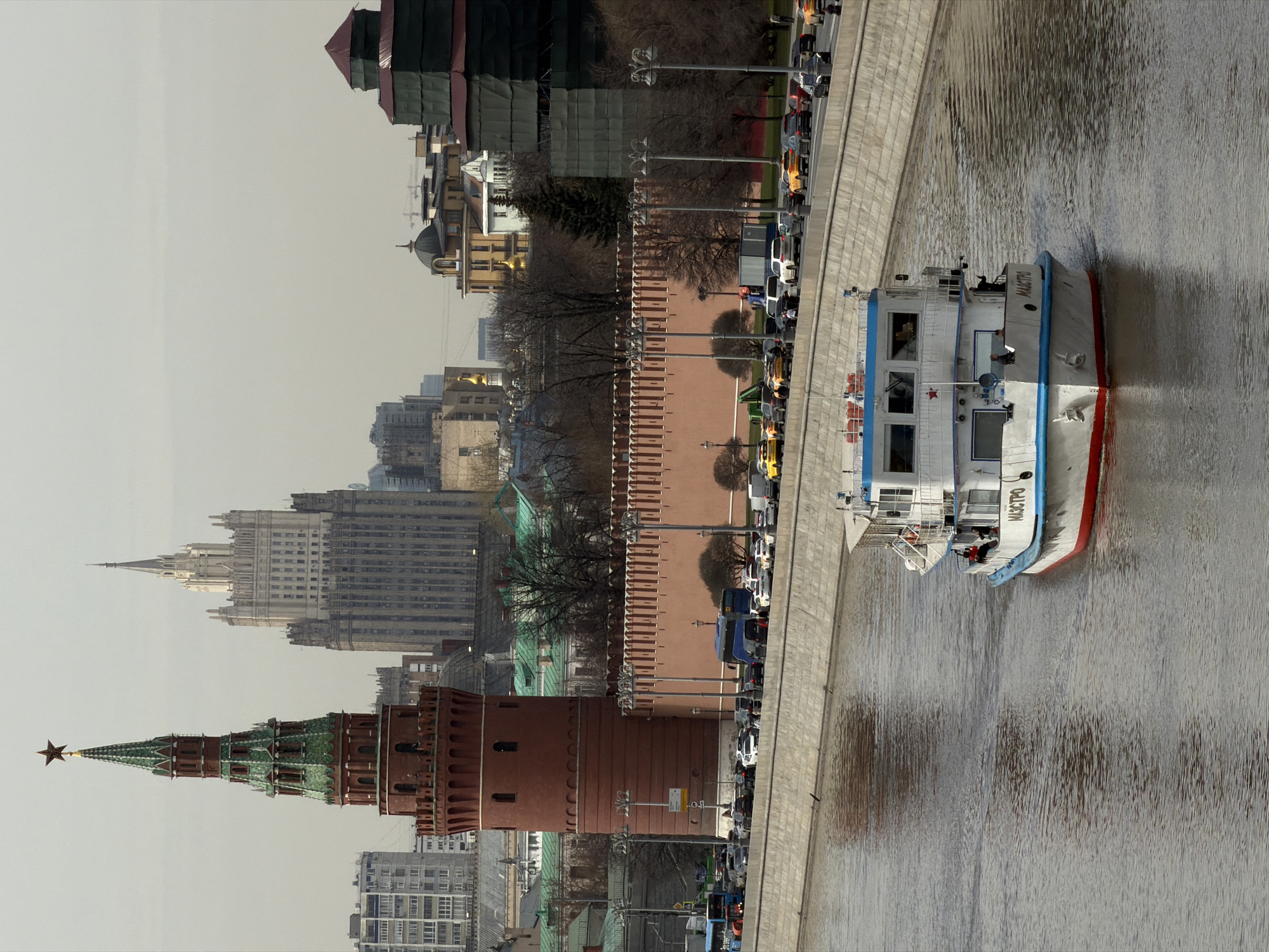 This image shows a cityscape in Moscow, Russia. A boat labeled "MAESTRO" is on the water to the right, and a concrete embankment runs along the water's edge, lined with numerous vehicles. The Kremlin walls and towers are visible in the center, alongside other buildings and structures, including a skyscraper in the background. The scene appears to be taken on an overcast day.