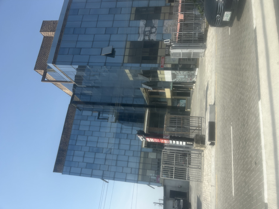 The image shows a modern glass and brick building in the city of Victoria Island, Nigeria, located at the coordinates latitude: 6.426, longitude: 3.452. The building is a multi-story structure, characterized by a prominent glass facade divided into numerous square panels, reflecting the surrounding environment. It has a brown brick roof with a metal frame. The entrance to the building is marked by a red sign post, and is secured by a metal gate. The building is positioned on a paved street, with a black car parked to the right of the image. The area surrounding the building is clean and well-maintained. The sky is clear blue with a few light clouds, reflecting the bright, sunny day. There are no people or objects present in the image.